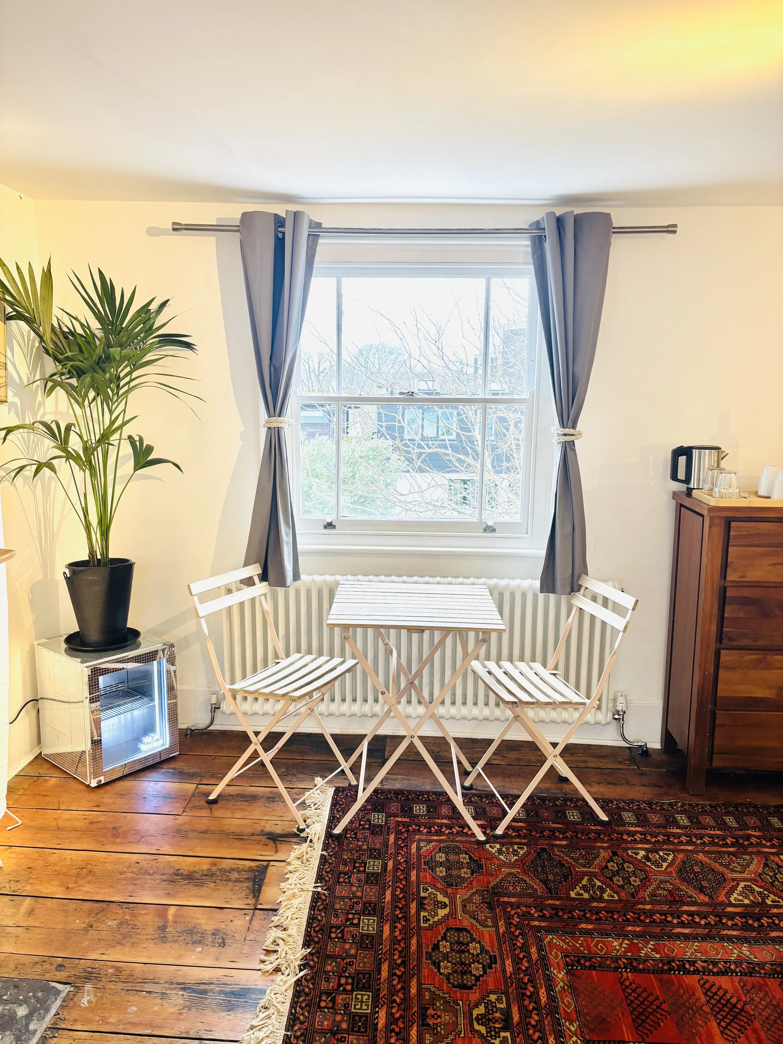 A cozy corner with a small white table and two matching folding chairs by a window with gray curtains tied back. A potted plant sits on a small metal side table, and a wooden cabinet with a kettle and glasses is nearby. A colorful patterned rug covers part of the wooden floor.