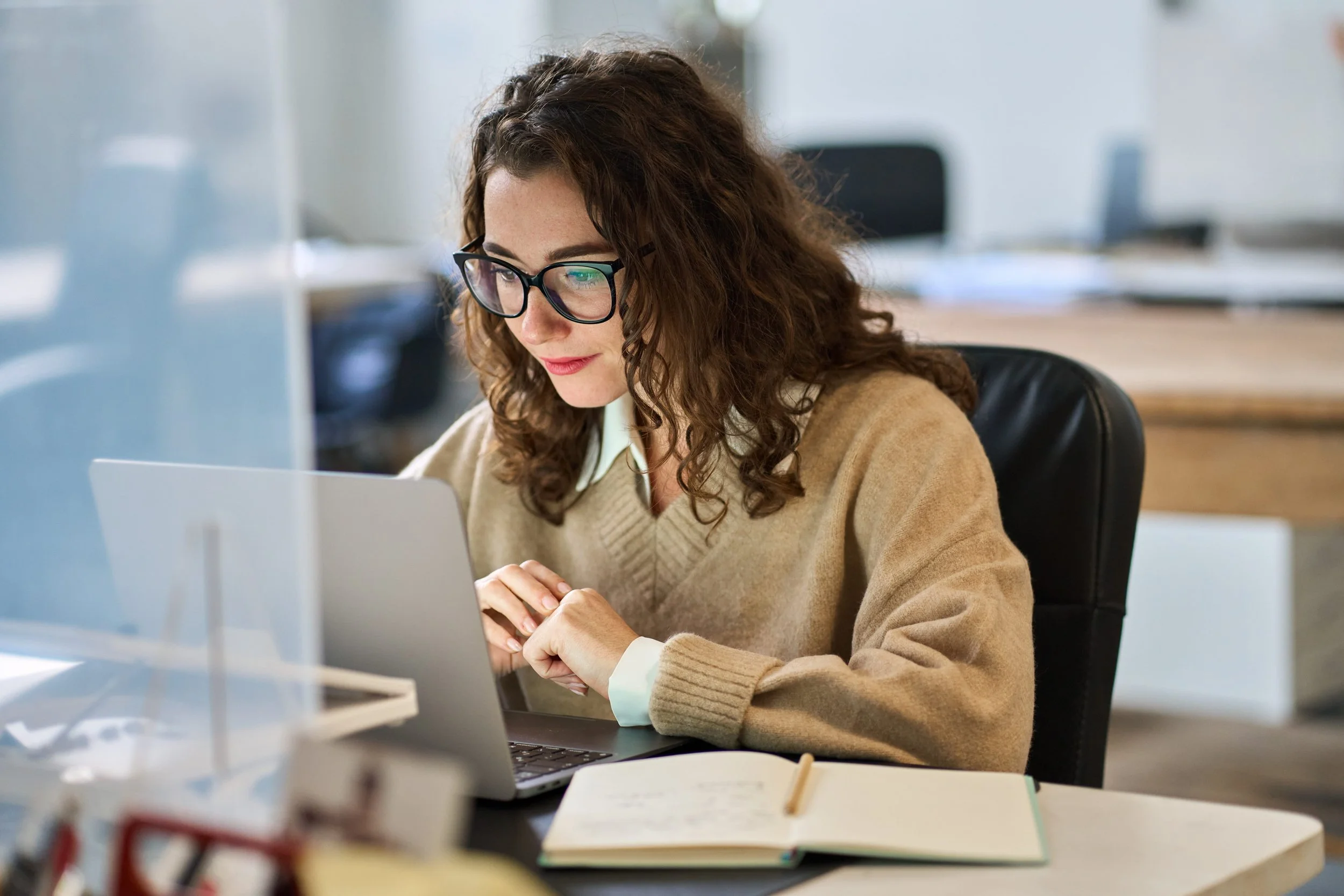 A woman with curly brown hair, wearing glasses, a beige sweater, and a white shirt, working at her computer in an office environment.