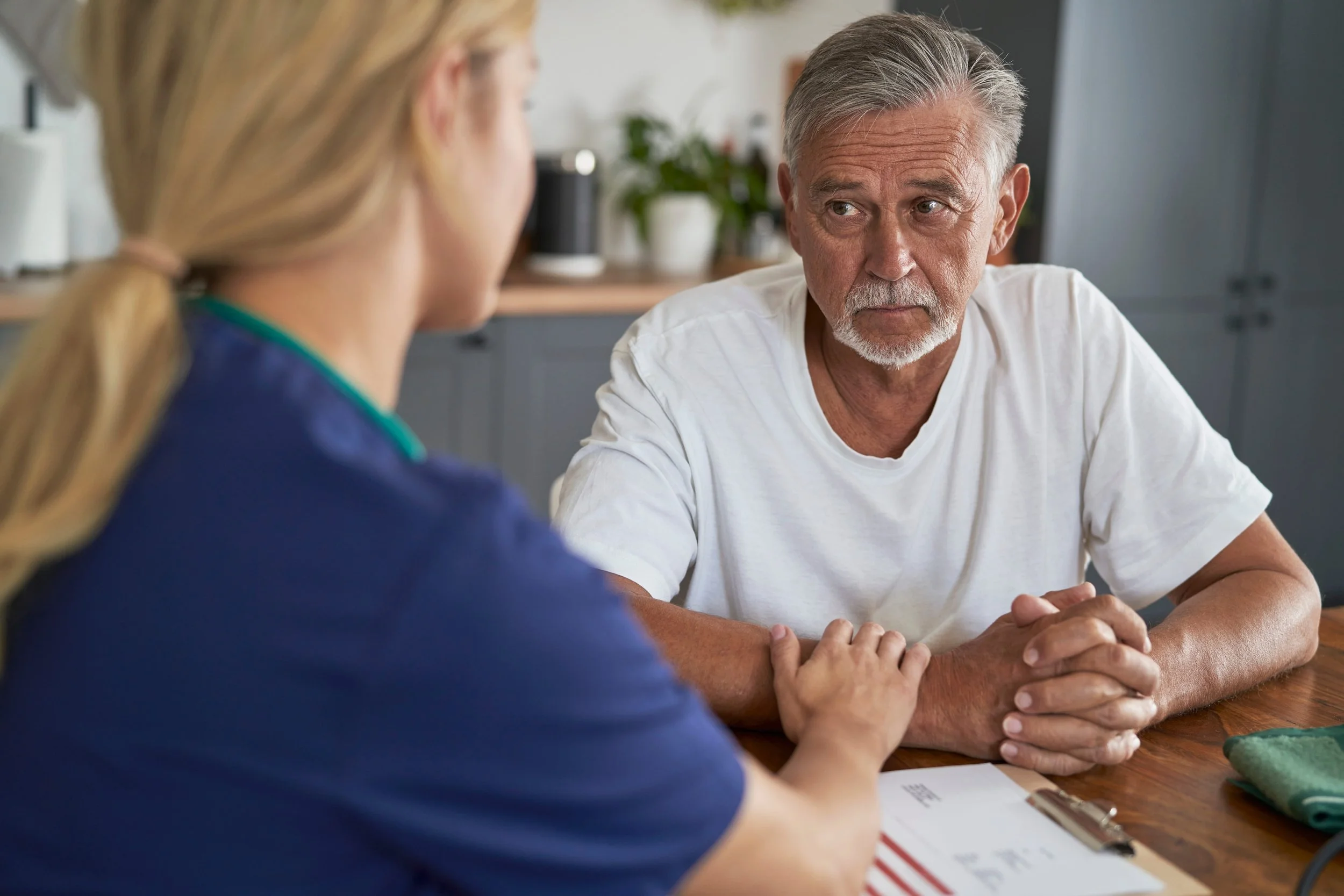An elderly man and a woman are sitting across from each other at a wooden table, engaging in a serious conversation. The man is looking attentively at the woman, with his hands resting on the table. The background shows a kitchen with some potted plants and a blurred wall.