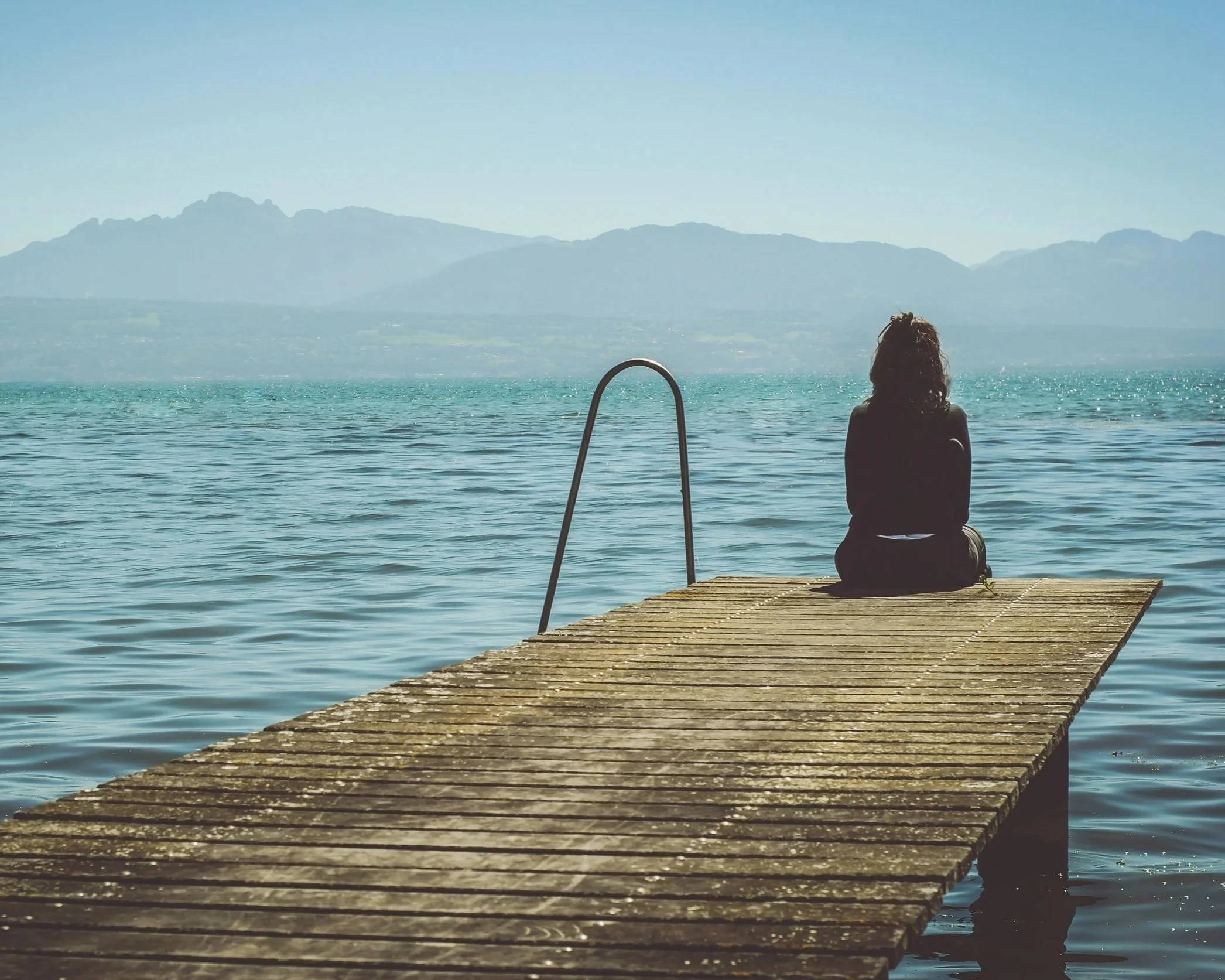 A person sitting on a wooden dock by a large body of water, looking toward distant mountains under a clear sky.