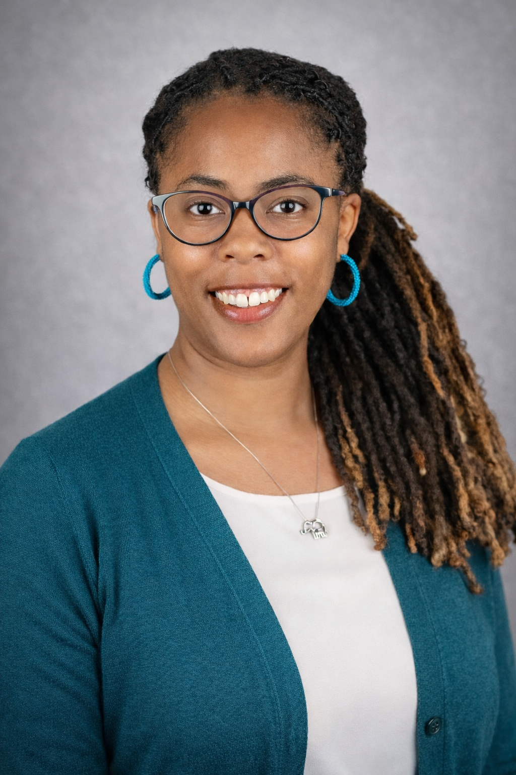 A woman with glasses and blue earrings smiling at the camera against a gray background.
