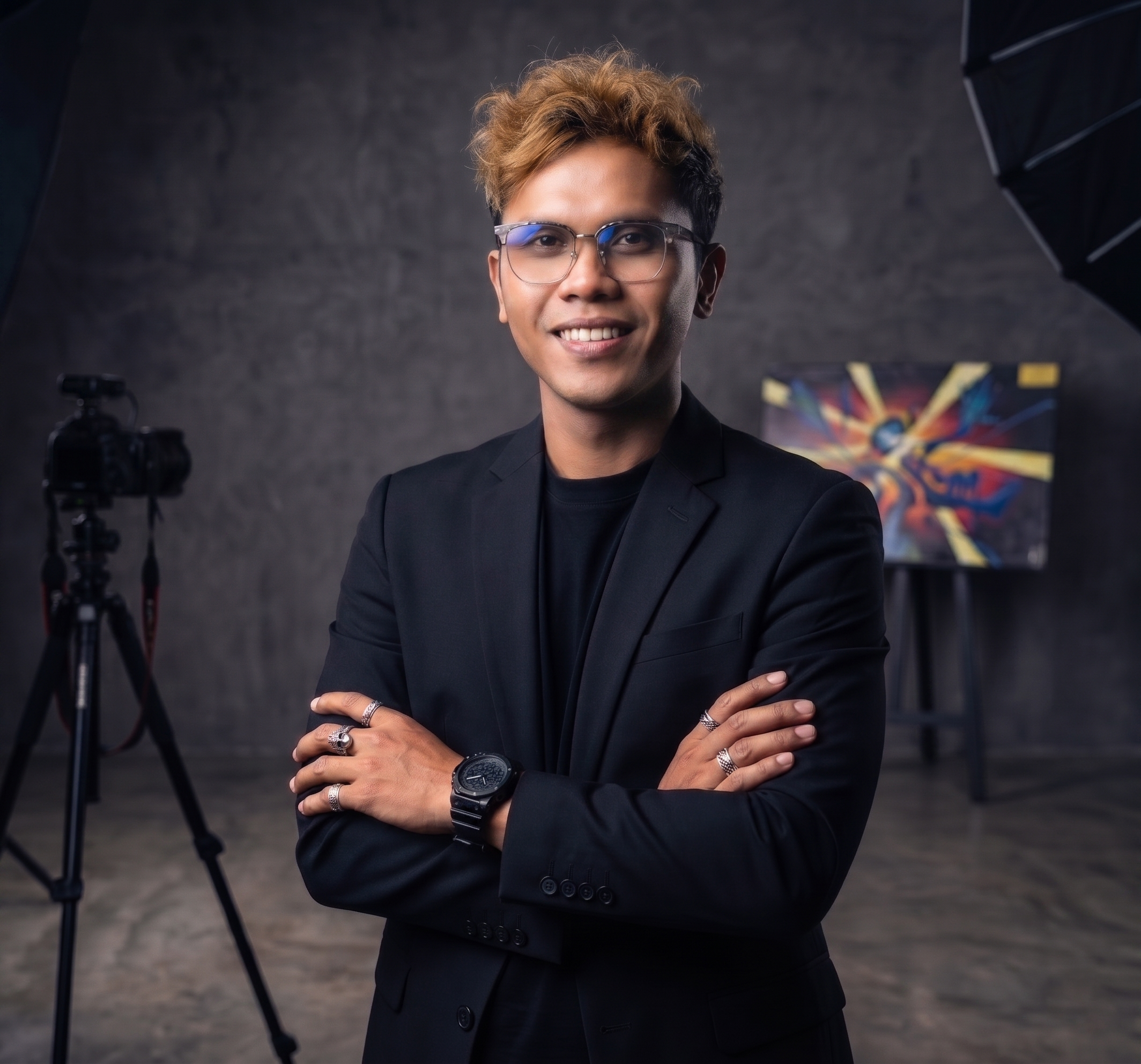 A young man with light skin, wearing glasses, a black blazer, and rings, smiling with arms crossed in a photography studio with professional lighting equipment and vibrant artwork in the background.