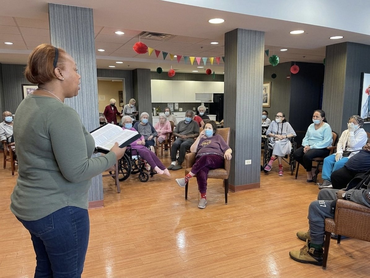 A woman stands in front of an audience reading from a book, with elderly people seated in a room decorated with colorful banners and hanging ornaments, all wearing face masks.