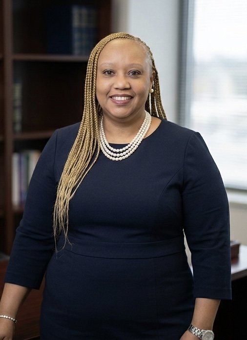 A smiling woman with long braided hair wearing a navy blue dress and pearl necklace standing in an office with bookshelves and a window in the background.