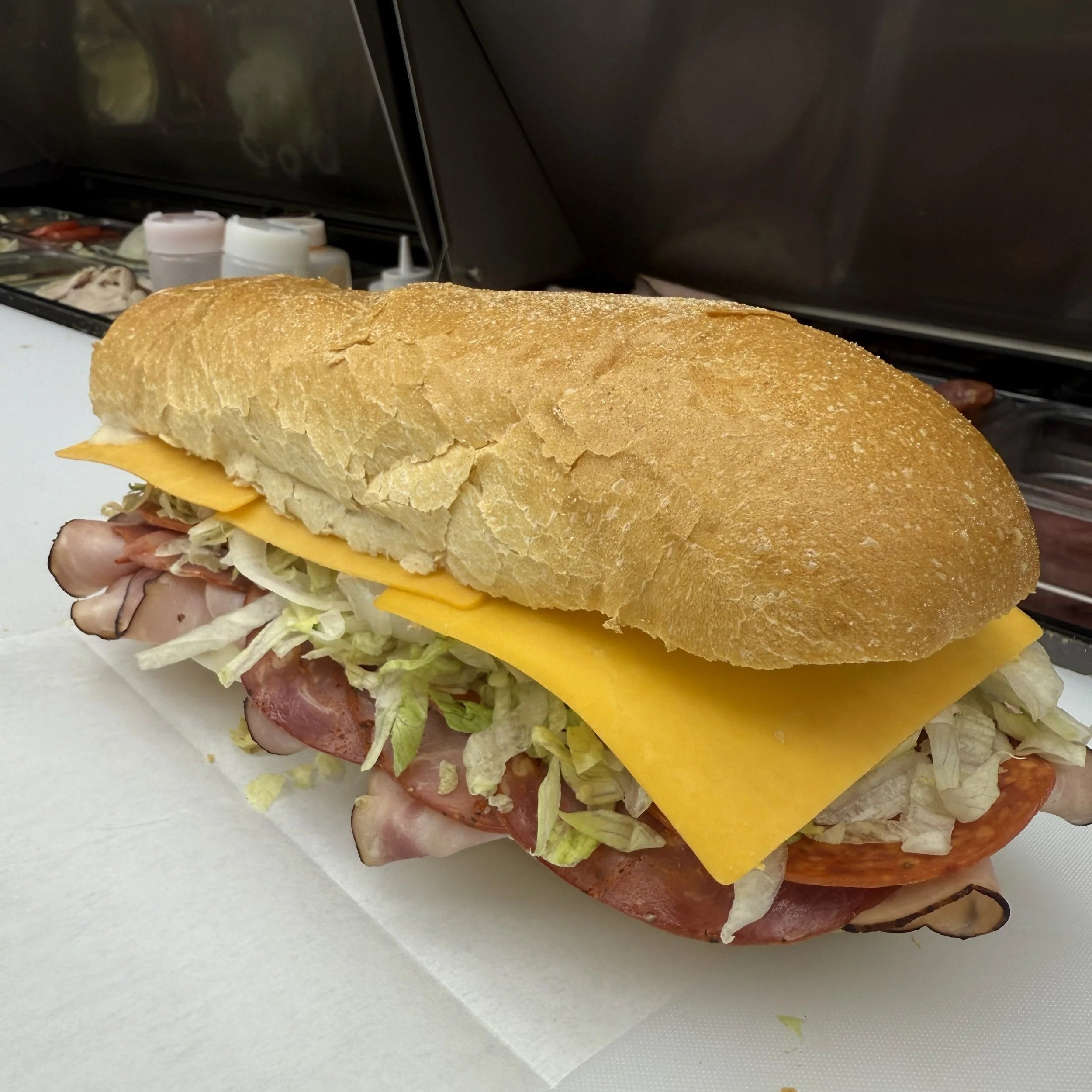 Close-up of a submarine sandwich with layers of deli meats, ham, cheddar cheese, lettuce, and a Pielies Bakery bun.