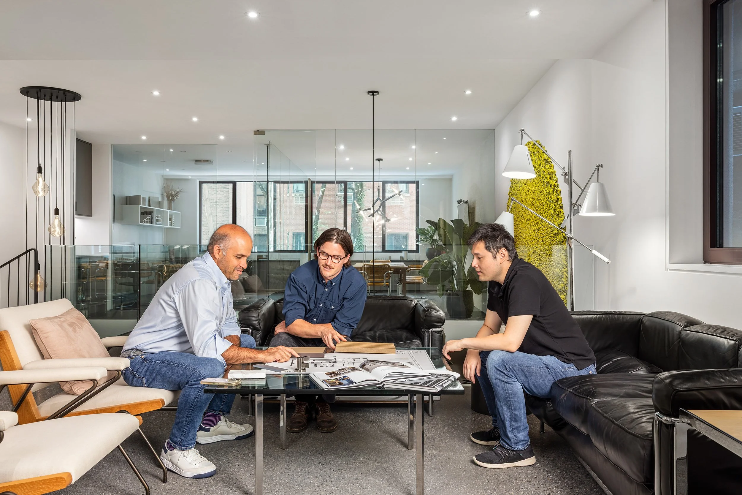 Three men sitting on black leather couches around a coffee table, engaged in a discussion with documents and a tablet on the table, in a modern, well-lit office space with large windows and decorative wall art.