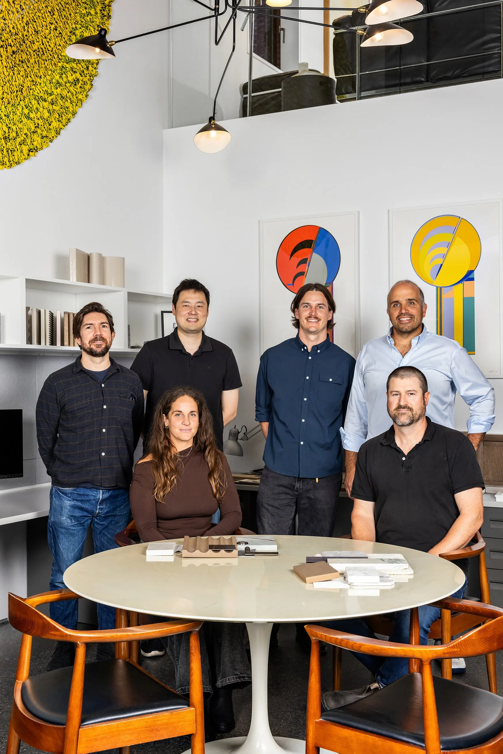 A group of eight diverse professionals, four standing and four sitting, in a modern office with white walls, bookshelf, and colorful abstract art. They are gathered around a white oval table with color samples on it, all smiling at the camera.