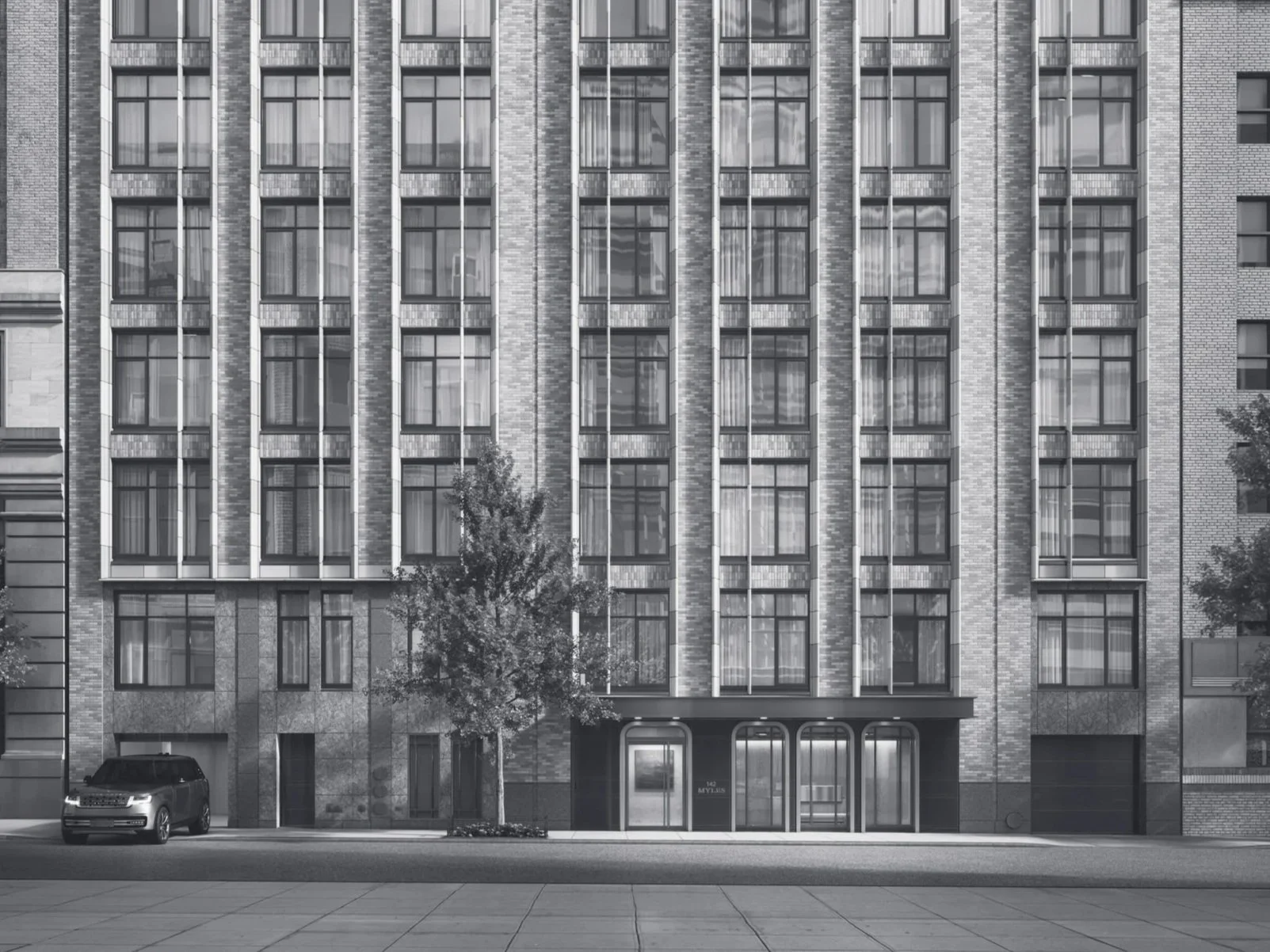 Front view of a multi-story modern apartment building with large glass windows, a tree in front, and a parked car on the street.