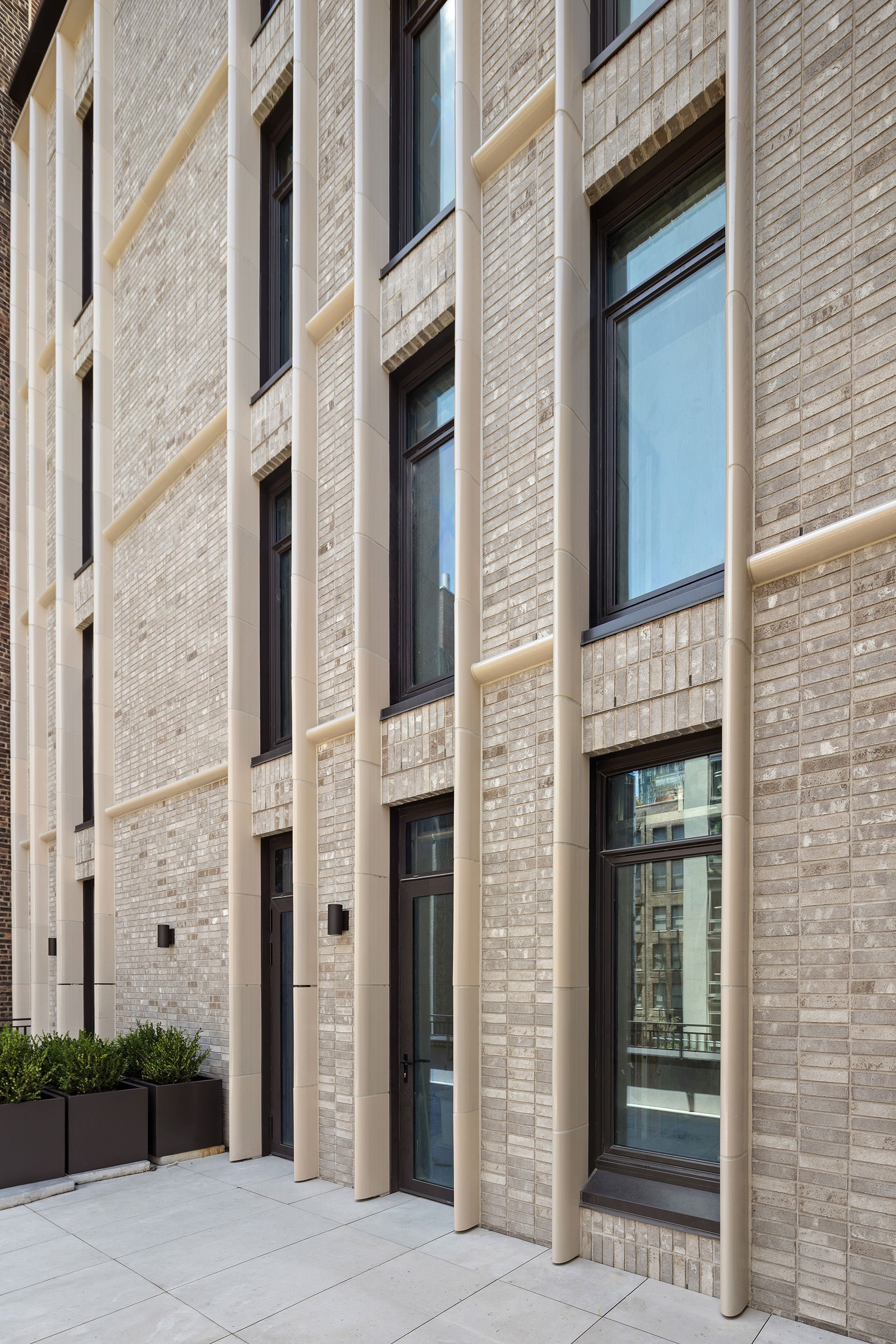 Modern residential building with beige brick facade, large black-framed windows, and decorative vertical tubes on the exterior. Potted plants are positioned at the base of the building.
