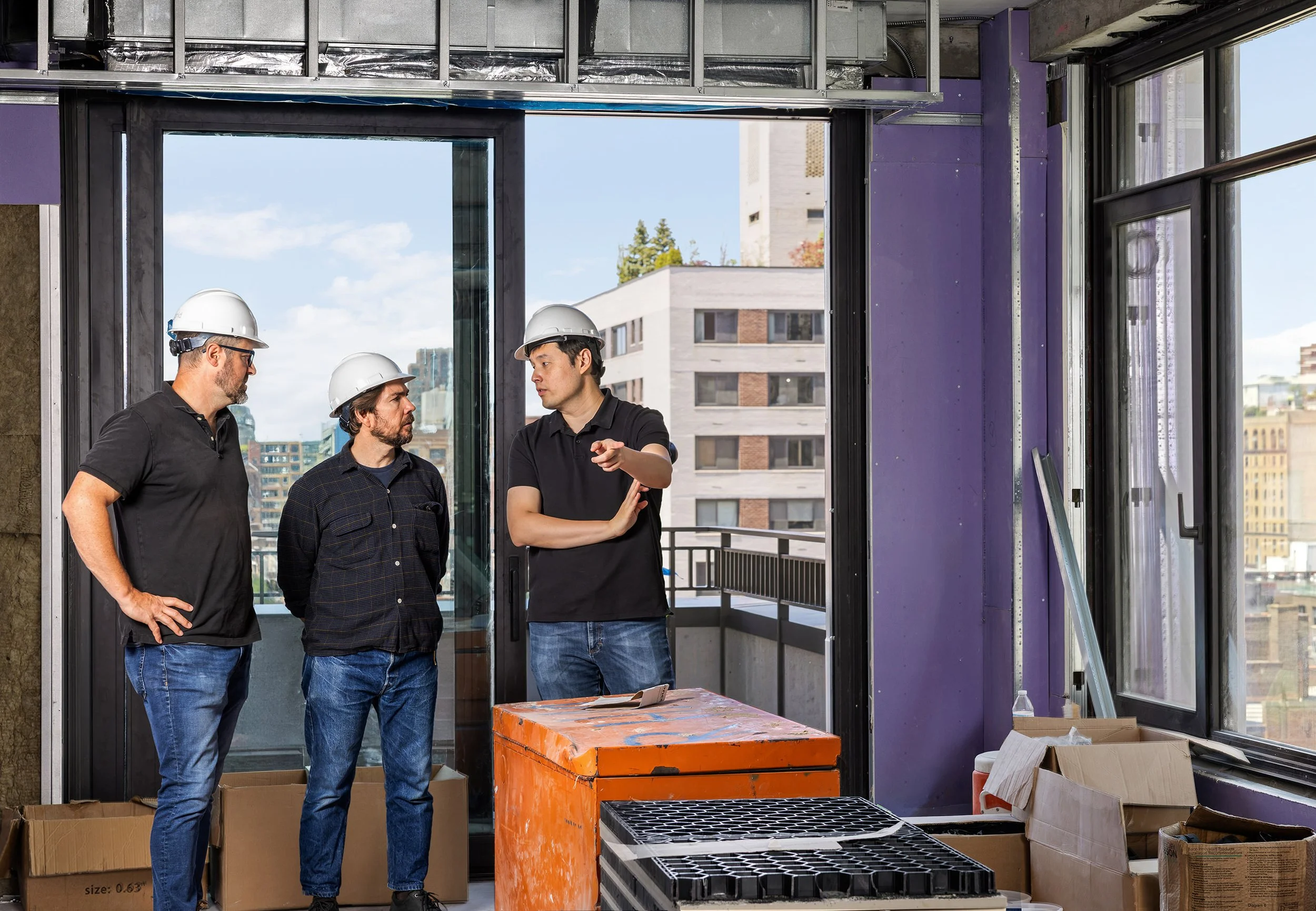 Three construction workers wearing hard hats and shirts, standing inside a partially built apartment with large windows overlooking city buildings, discussing construction plans.