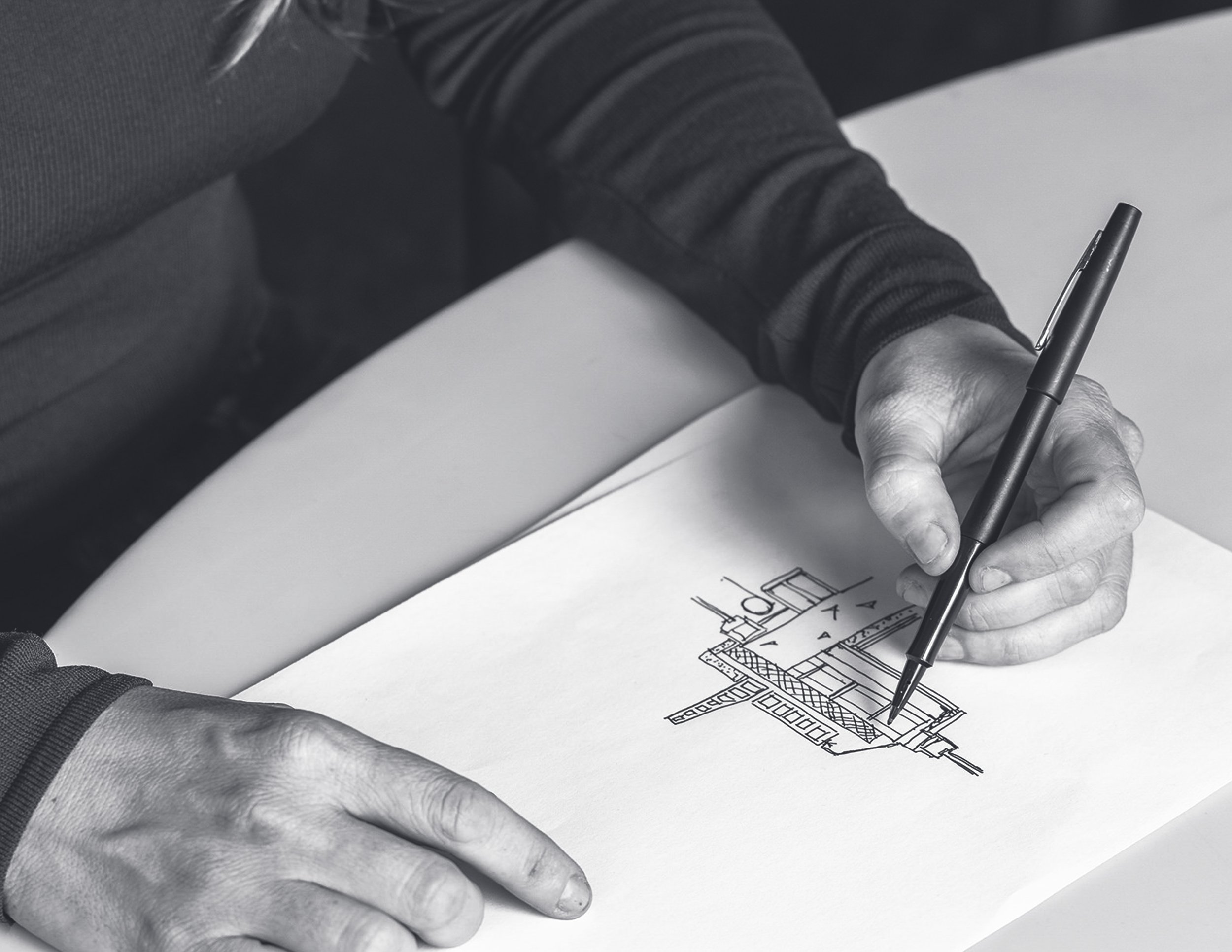 Black and white photograph of a person’s hands drawing a building blueprint or architectural plan on a sheet of paper with a black pen.
