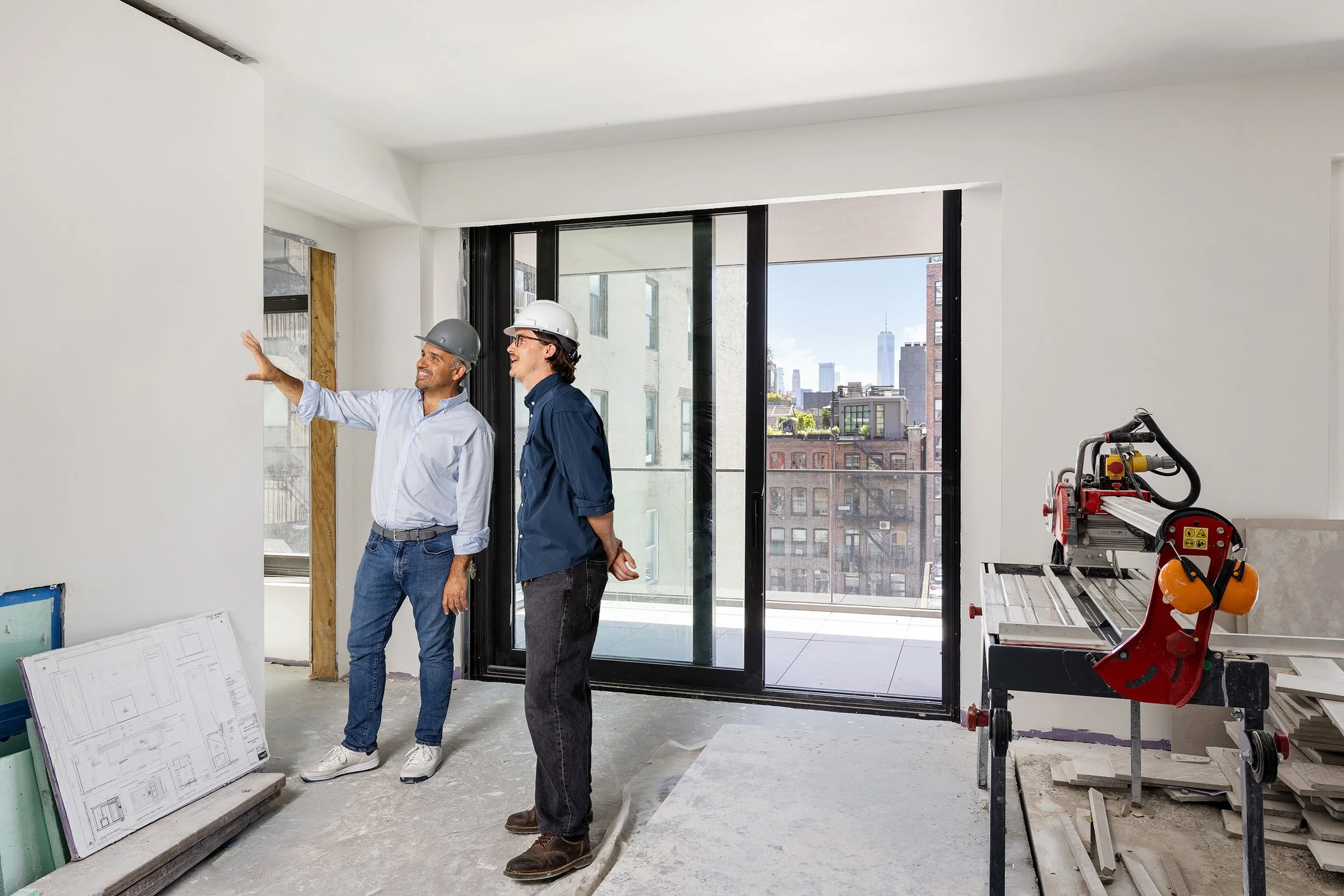 Two men wearing hard hats and casual clothing are inside a building under construction, having a conversation. There are construction materials and plans on the floor, large windows with a city skyline in the background, and construction equipment ne