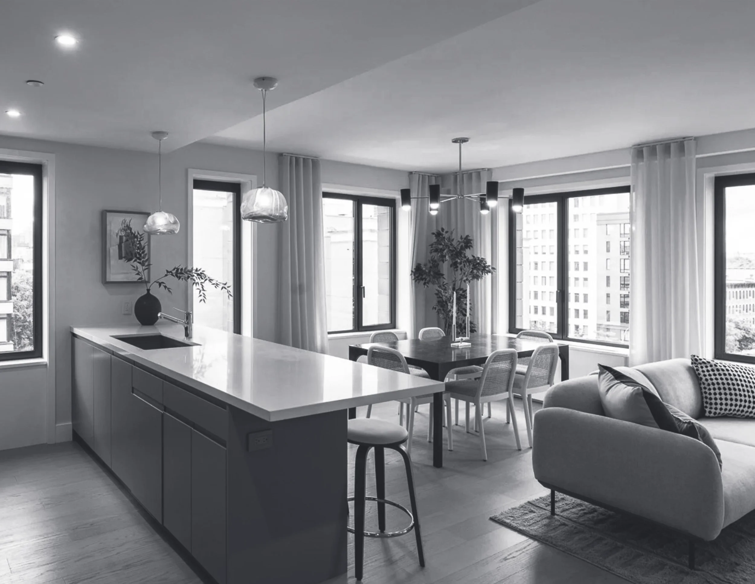 Modern apartment interior with large windows, a kitchen countertop, pendant lights, a dining table with chairs, and a couch near a rug and decorative pillows.