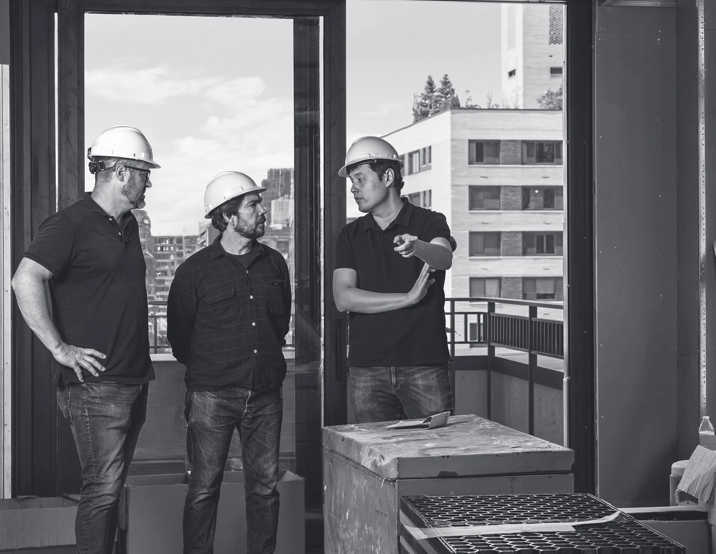 Three construction workers wearing safety helmets standing inside a building, engaged in a discussion, with a cityscape outside the window.