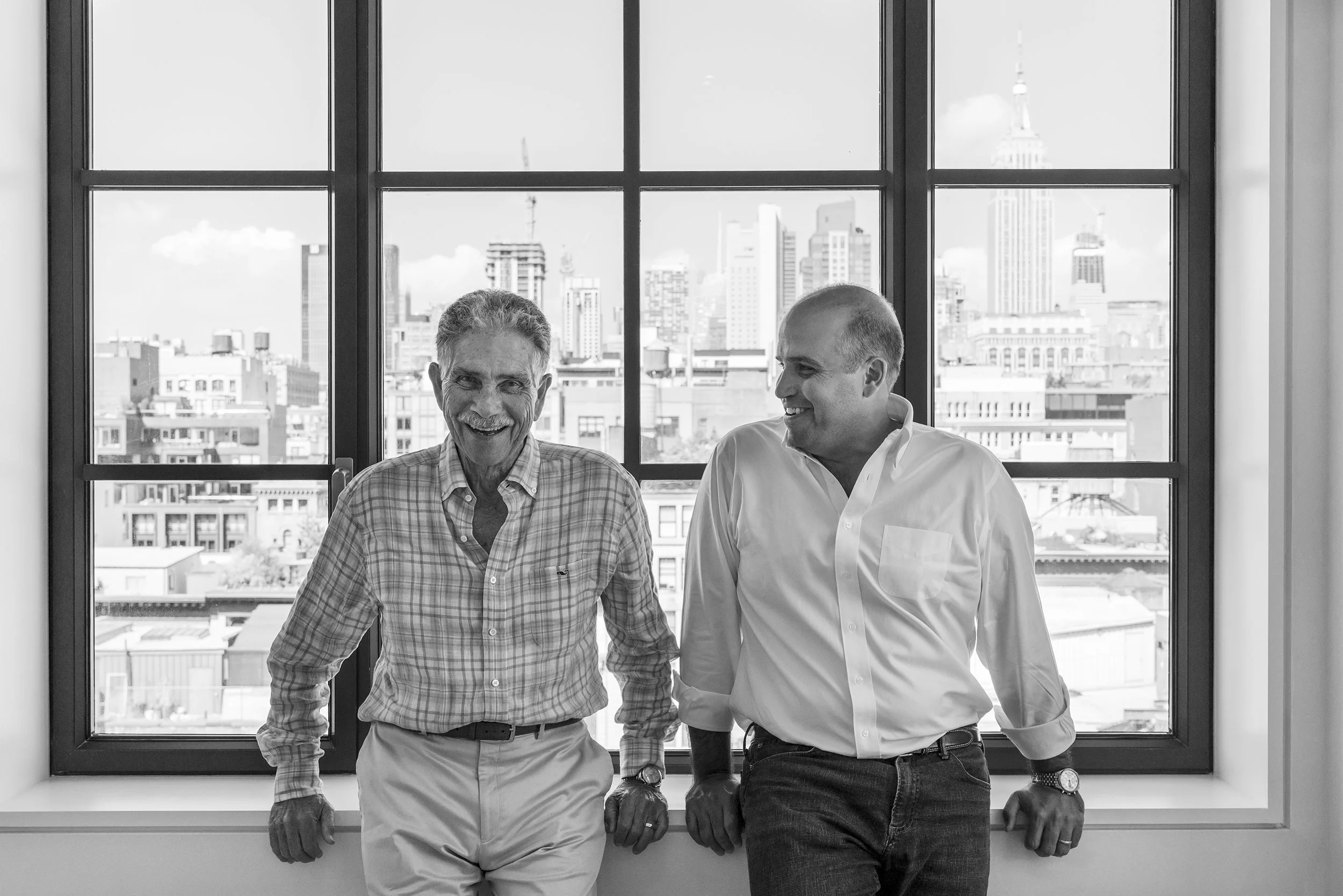 Two men are sitting by a large window with a city skyline in the background. They are smiling and appear to be having a conversation.