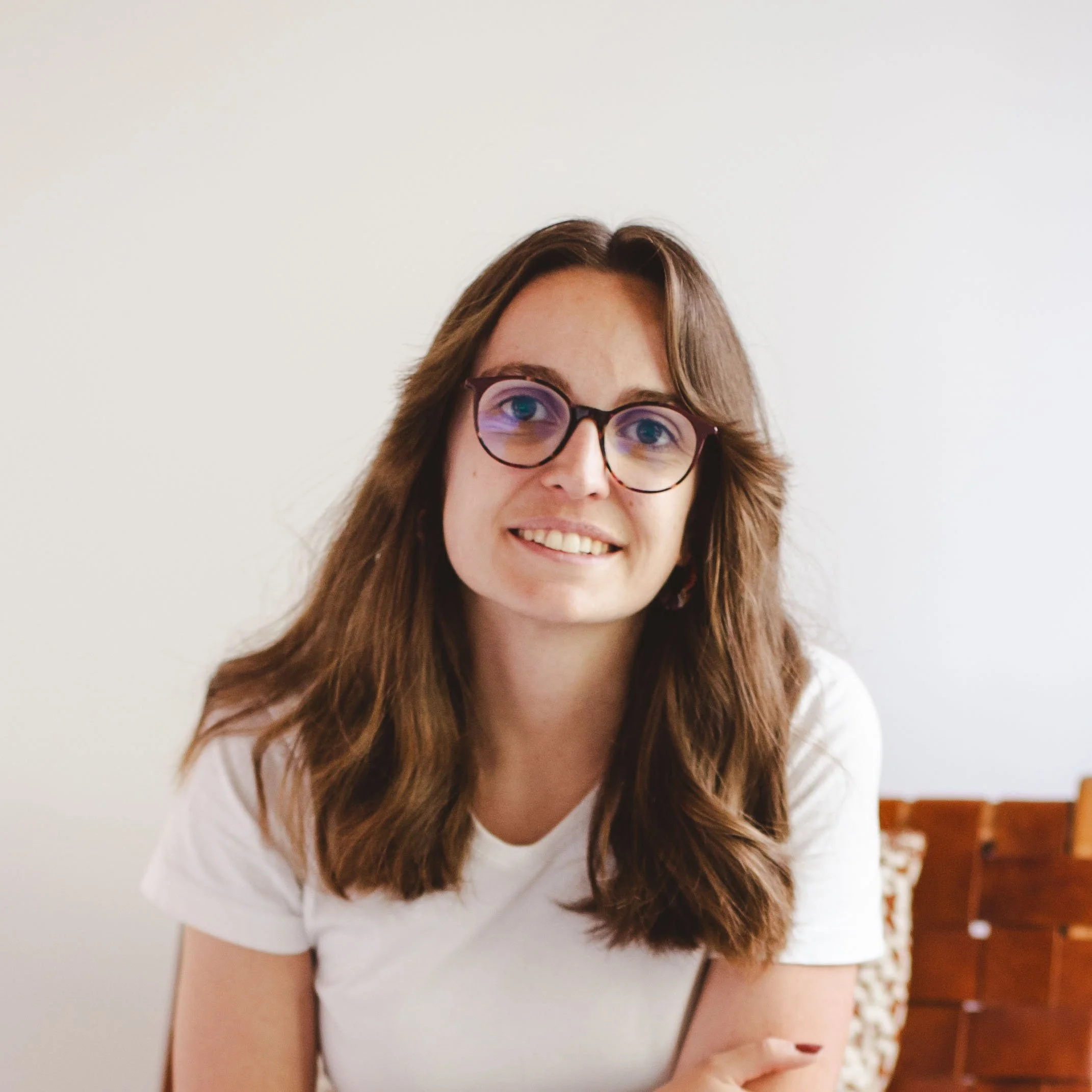 Une femme avec des cheveux longs et bruns porte des lunettes rondes, souriant légèrement, vêtue d'un t-shirt blanc, dans un intérieur sur fond clair