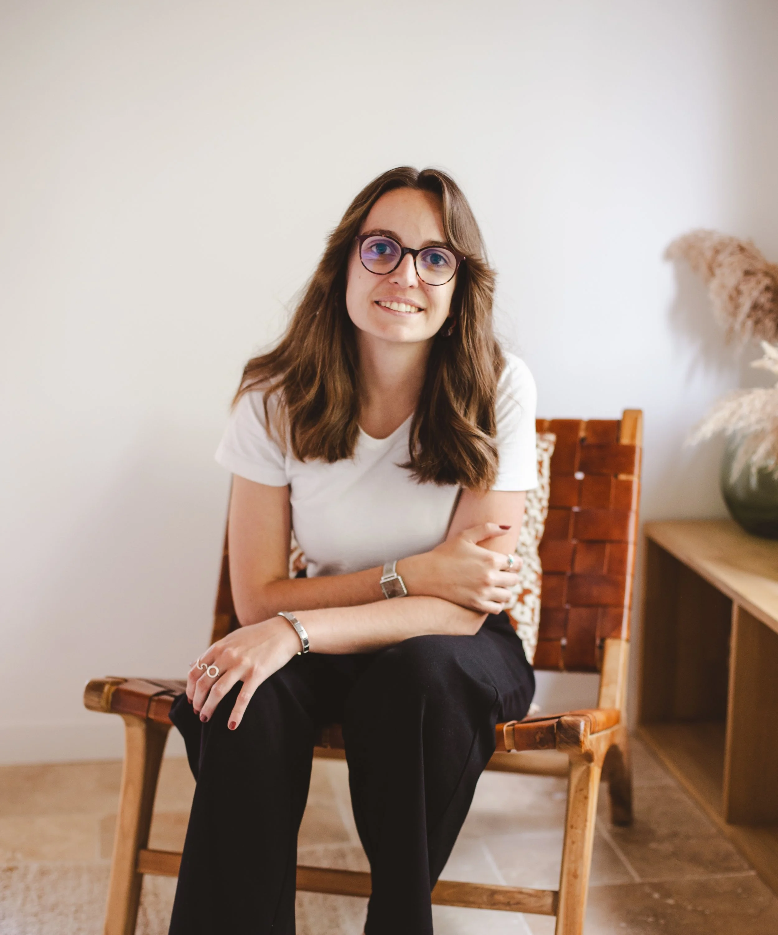 Une femme assise sur une chaise en bois, souriante, portant des lunettes rondes, un t-shirt blanc, avec un décor minimaliste et un grand vase à droite.