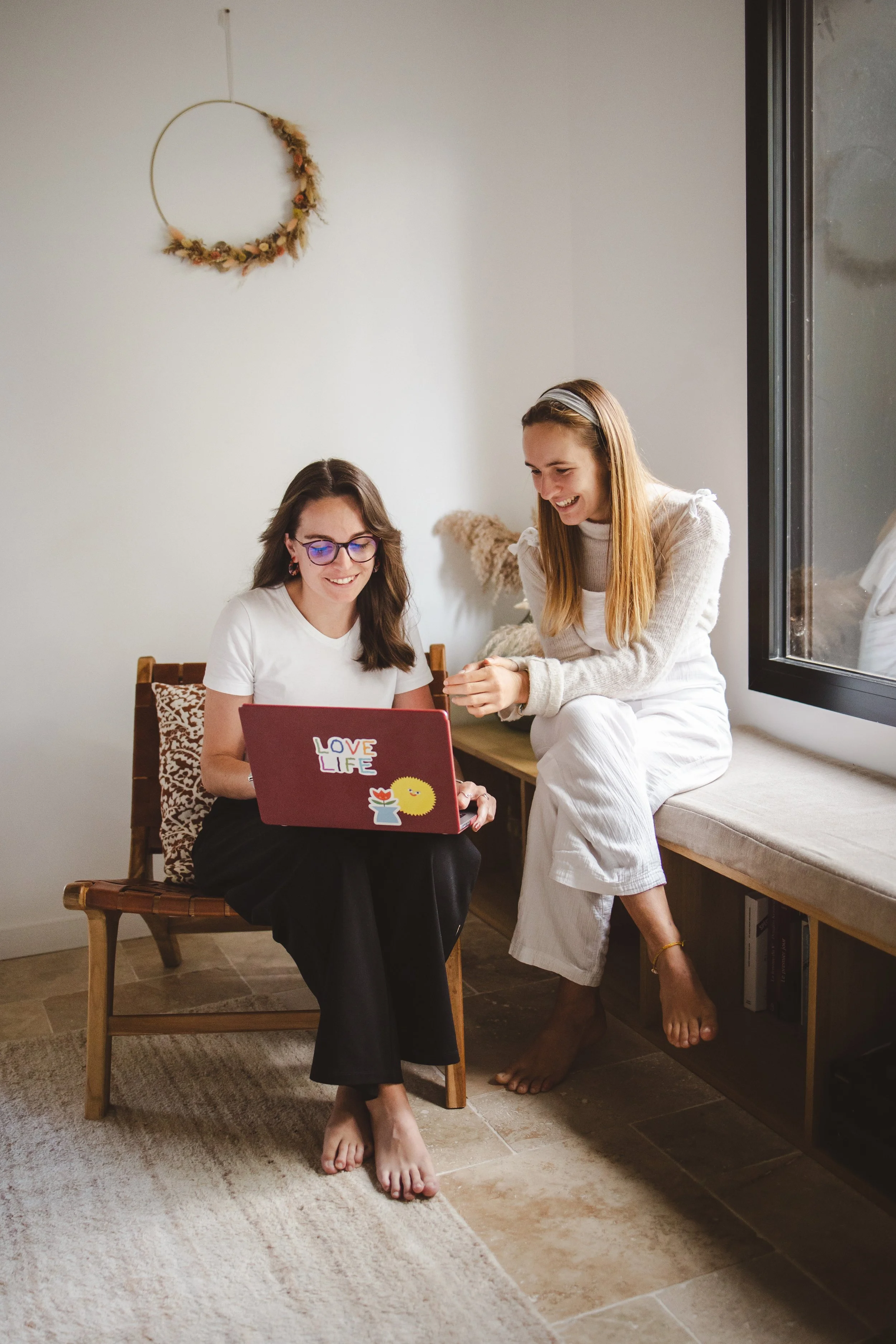 Deux jeunes femmes souriantes regardent un ordinateur portable dans une pièce lumineuse. L'une est assise sur une chaise en bois, portant des lunettes et un t-shirt blanc, l'autre est debout à côté d'elle, vêtue d'une robe blanche, les pieds nus sur le sol. Il y a une fenêtre à droite, un mur blanc avec une décoration florale suspendue, et une étagère avec des livres en dessous de la fenêtre.