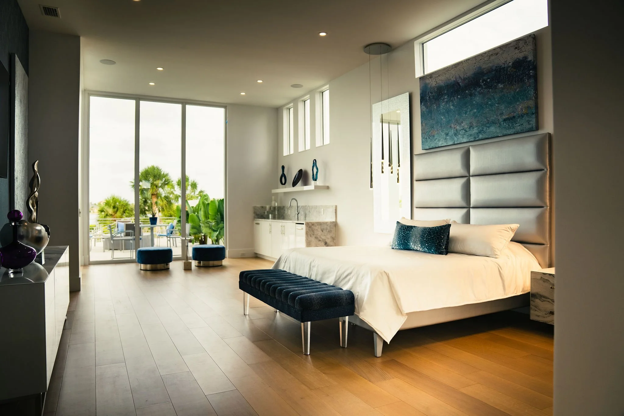 Modern hotel room with a large bed, blue pillows, wooden floor, artwork on the wall, and a balcony with outdoor seating and palm trees visible through sliding glass doors.