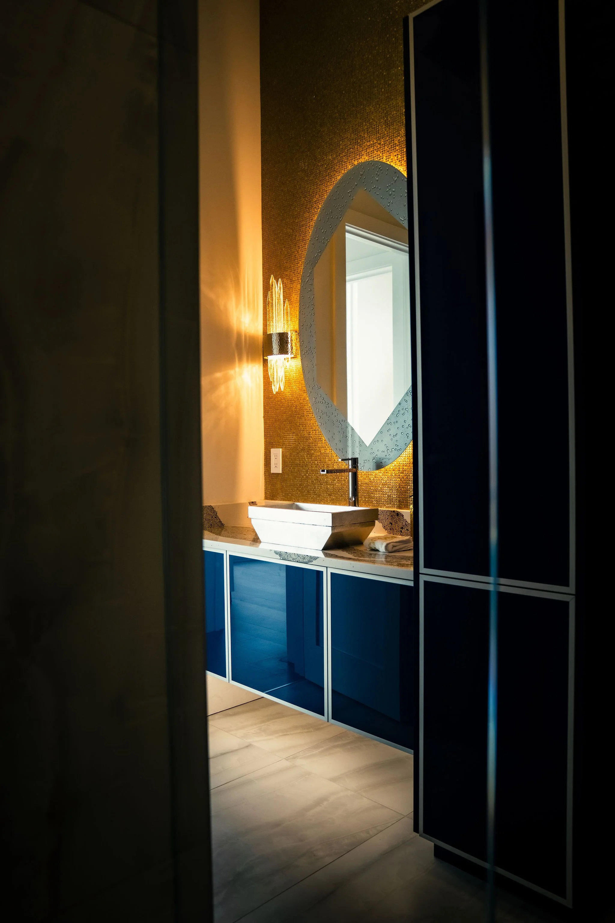 Interior view of a modern bathroom with a vessel sink, a geometric mirror, and blue cabinetry, partially viewed through a doorway with warm lighting.