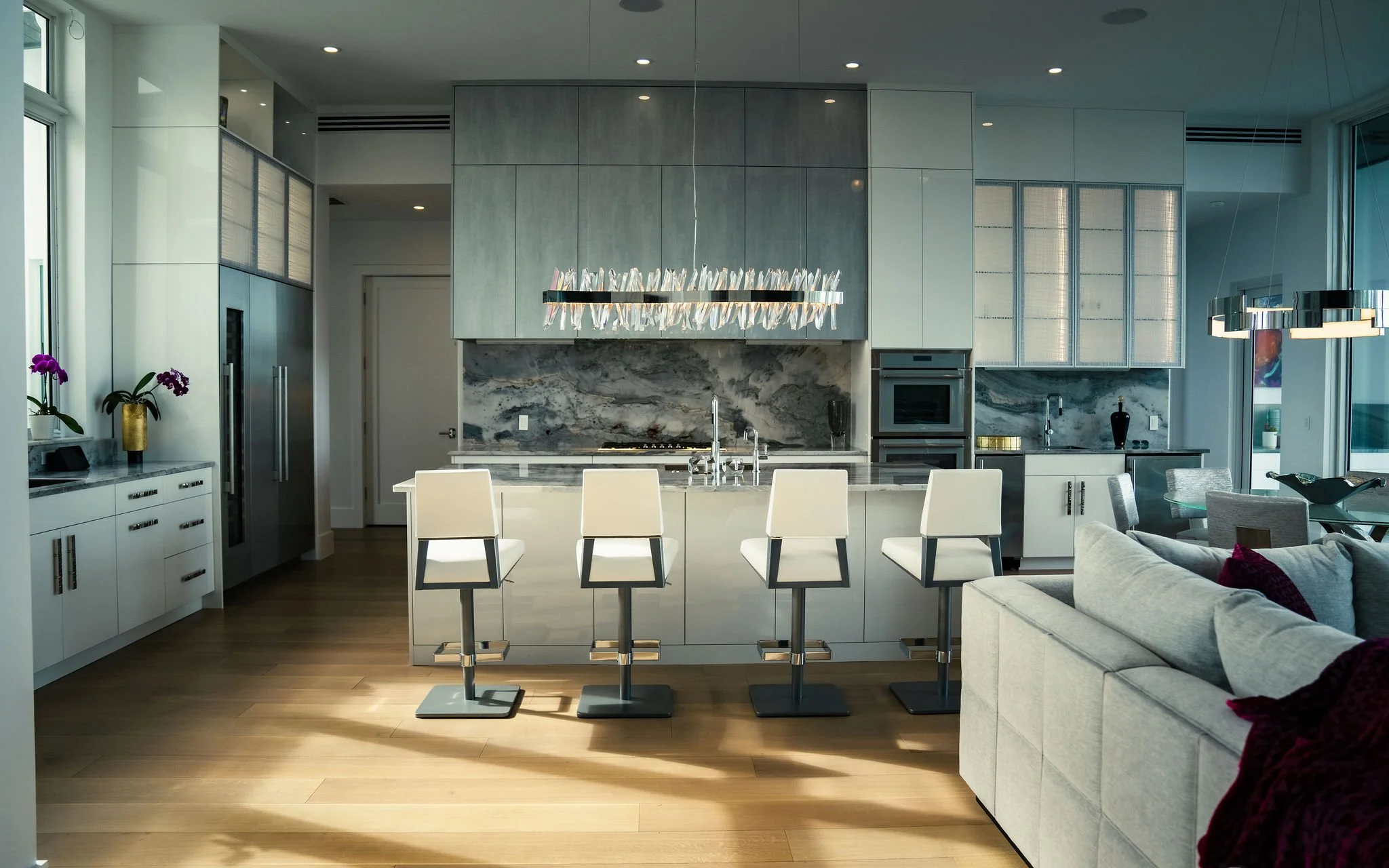 Modern kitchen with white cabinetry, marble backsplash, island with four white chairs, stainless steel appliances, and contemporary lighting fixtures.
