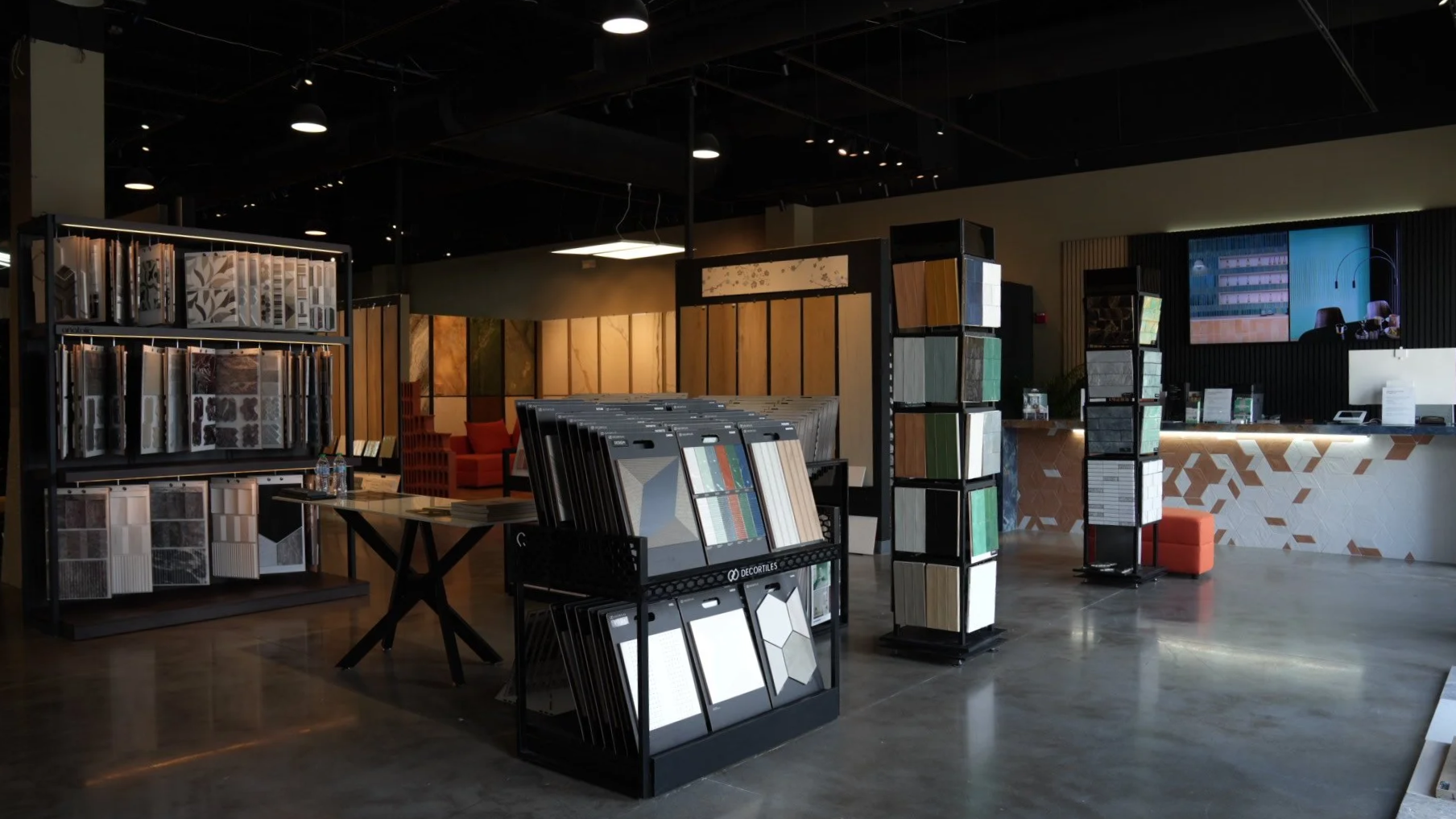 Interior view of a showrooms with display racks of tiles and flooring samples, a front desk, and a television screen on the wall.