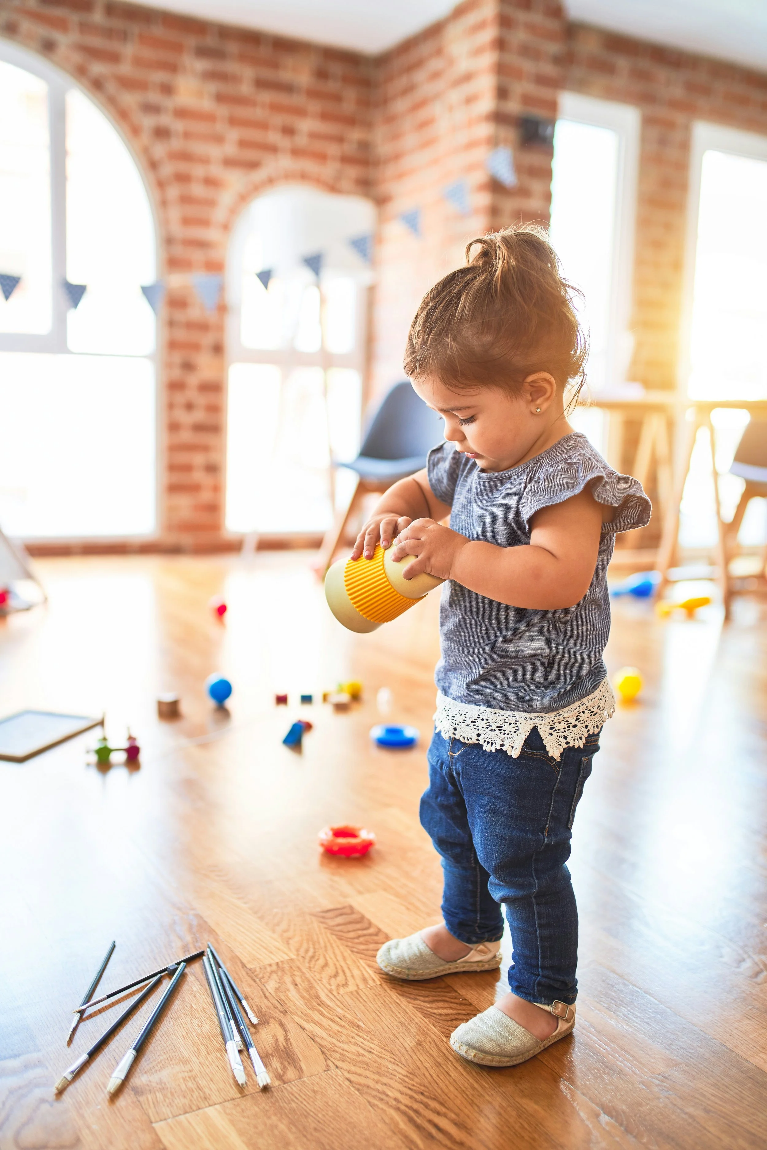 A young girl with brown hair in a ponytail, wearing a gray shirt with ruffled sleeves, dark blue jeans, and beige slip-on shoes, is standing on a wooden floor in a room with brick walls and large windows. She is playing with a yellow and orange toy, and there are scattered toys and black sticks on the floor around her.