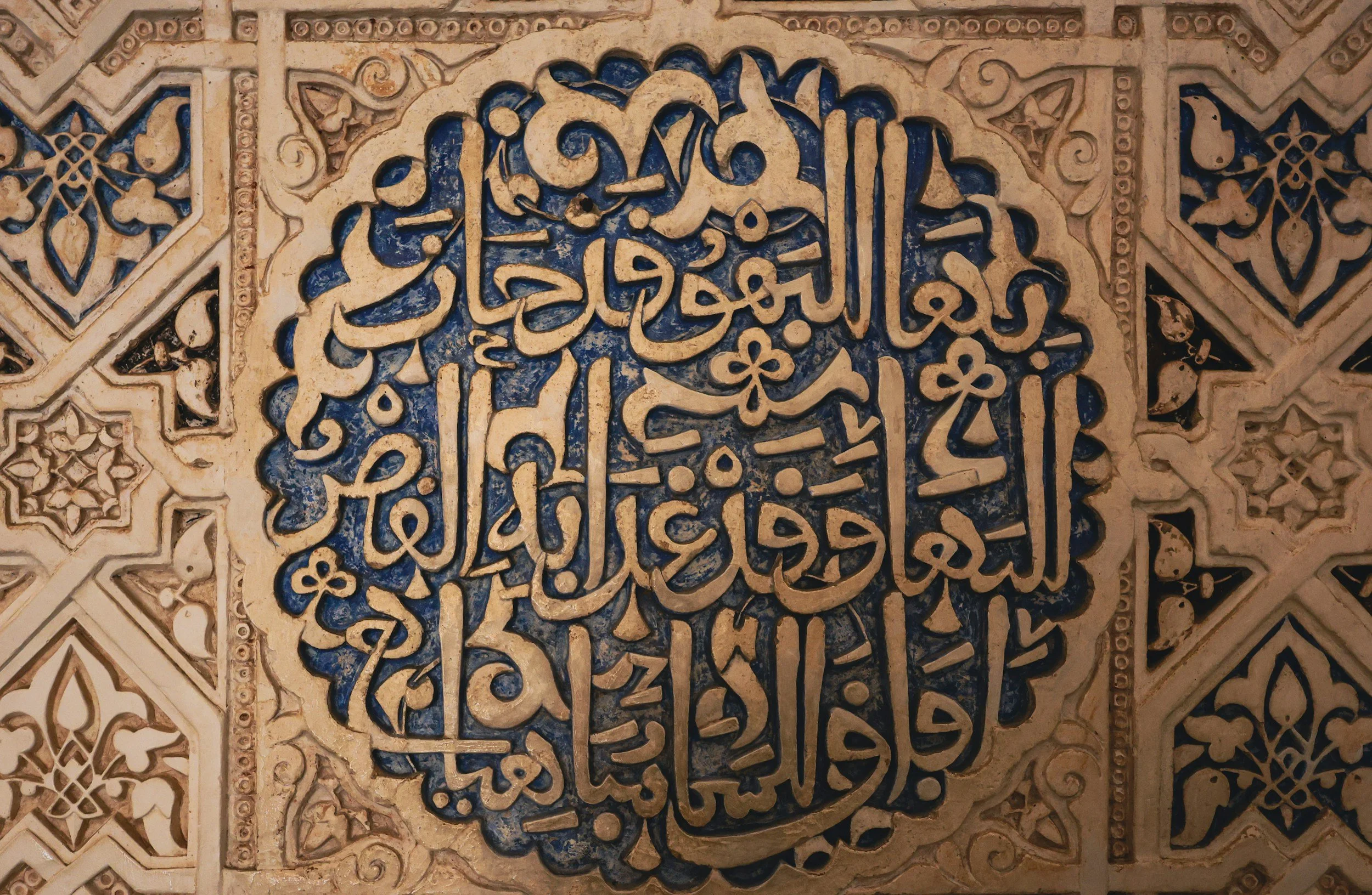 Close-up of a decorative Islamic calligraphy panel with intricate Arabic script in cream, surrounded by geometric patterns and floral motifs, with a blue background.