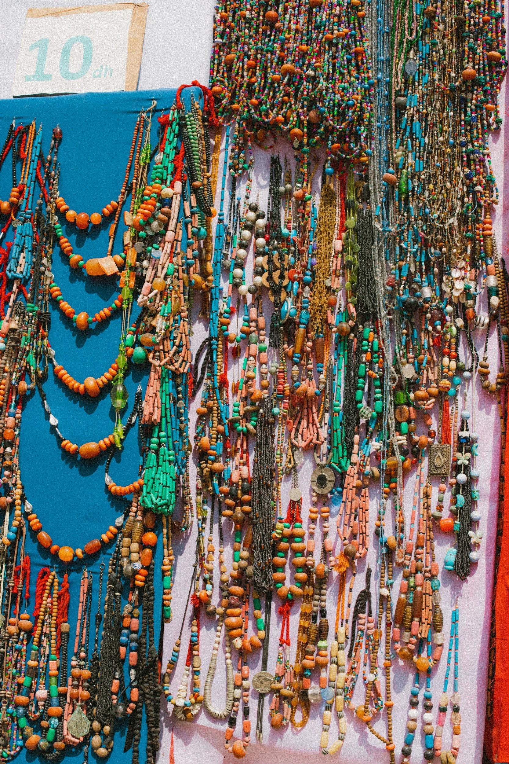 A display of colorful beaded necklaces hanging on a wall, with a white sign showing the price of 10 dh in the background.