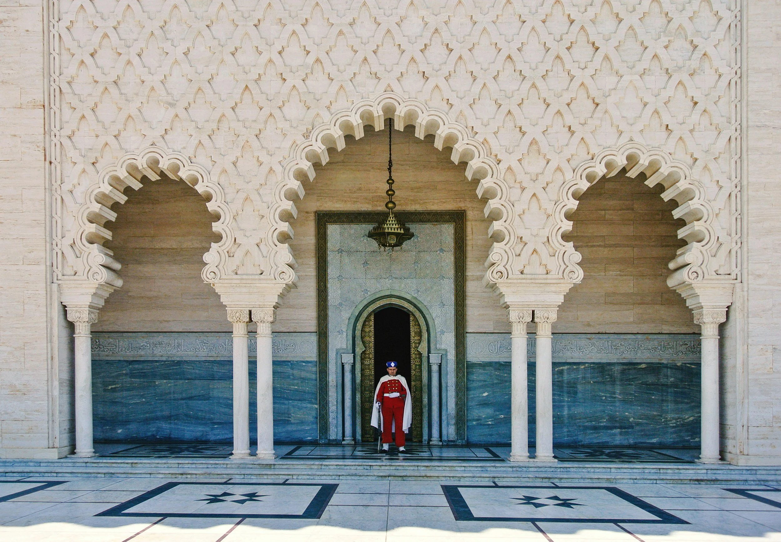 A large, ornate architectural entrance with three arches featuring intricate geometric patterns, supported by slender columns. A guard in a red uniform stands at the center beneath the middle arch, with a hanging lamp above. The floor has a decorative black and white geometric pattern.