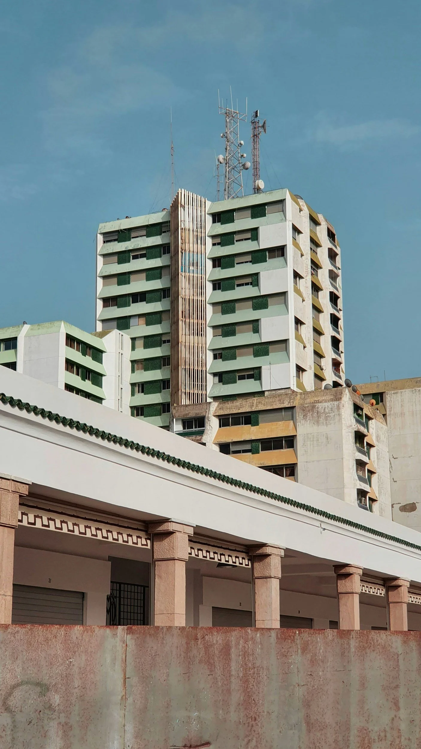 A multi-story building with antennas on the roof, featuring green balconies and white exterior walls. Some wear and tear are visible, and there is a lower structure with columns in the foreground.