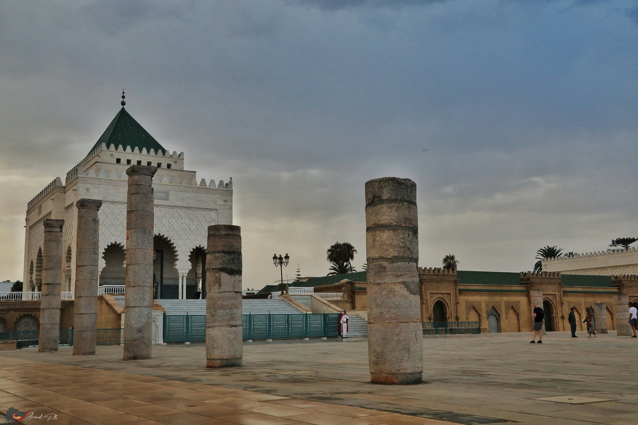 Historical site with ancient columns and a white building with a green roof, under a cloudy sky, with visitors walking around.