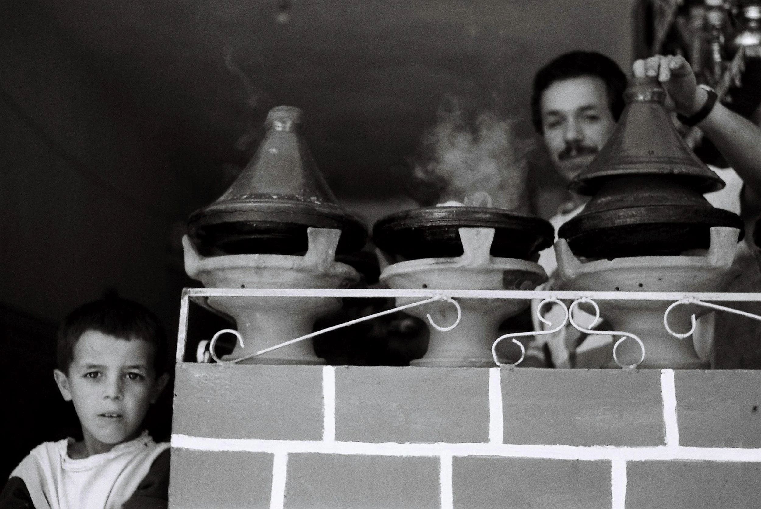 A black and white photo of a young boy with dark hair and expressive eyes, standing in front of a shelf with ceramic vases. A man is partially visible in the background, looking through the vases.