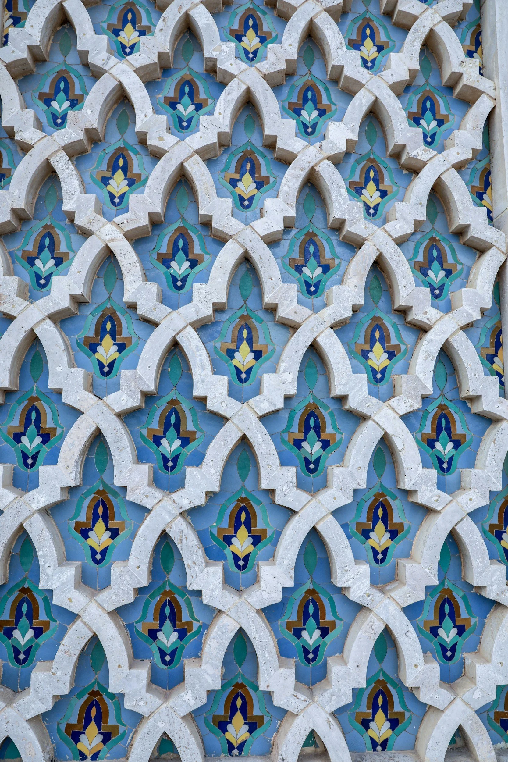 Close-up of an intricate mosaic tile wall with geometric and floral patterns in blue, turquoise, yellow, and brown, framed by white stone arches.