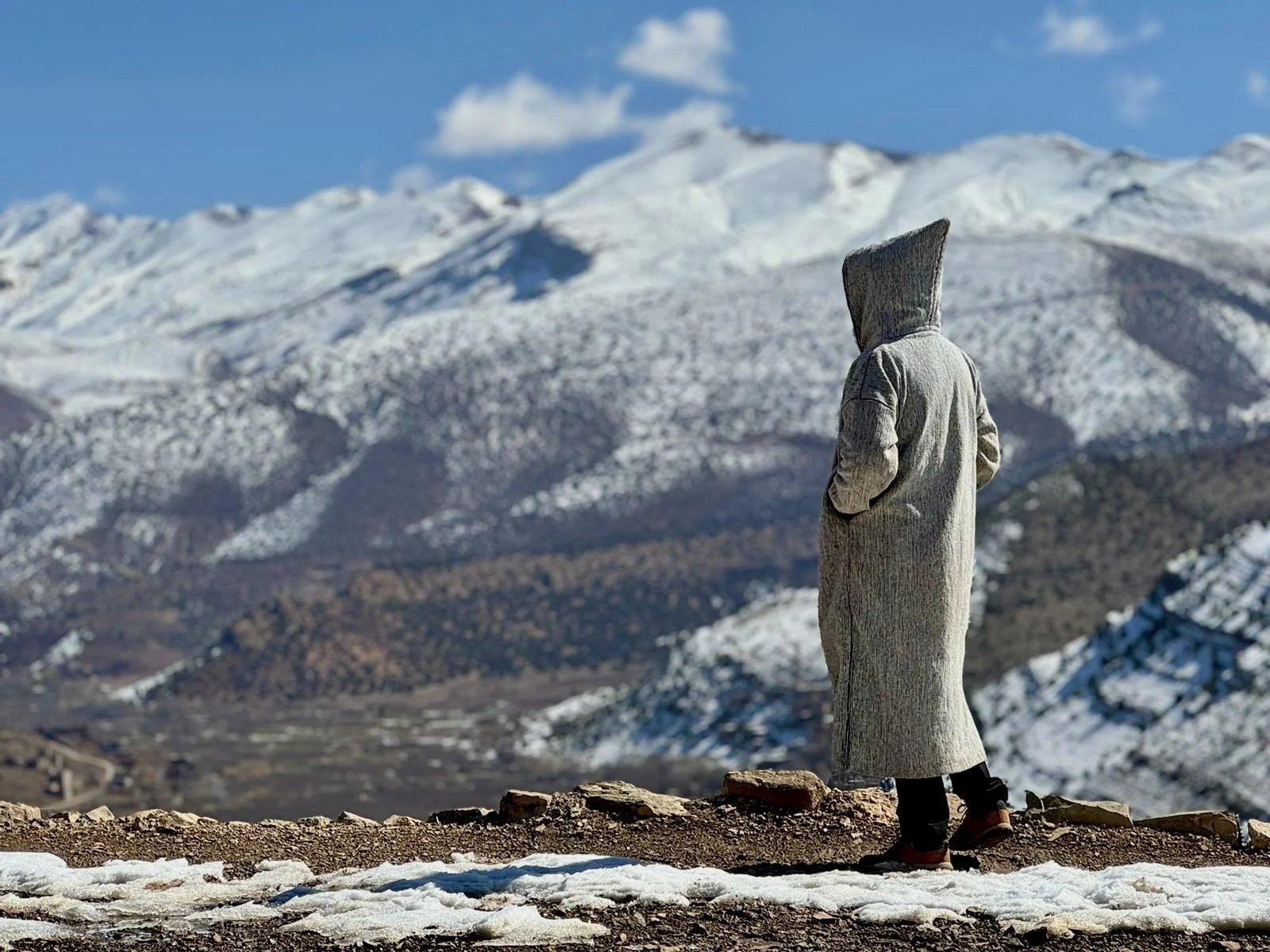 Person with a hoodie and coat standing on a mountaintop in winter, looking at snow-covered mountains under a blue sky.