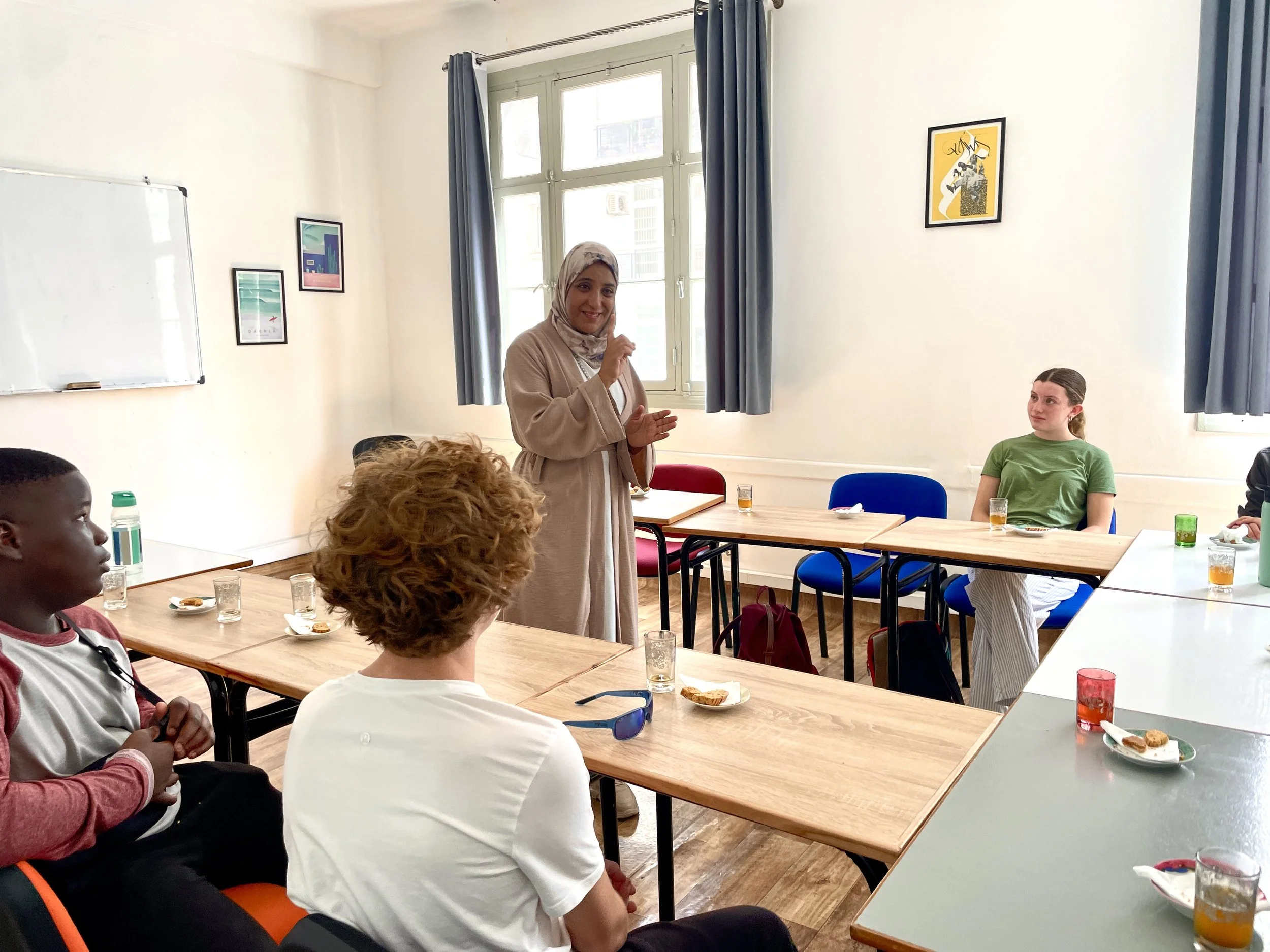 A woman in a beige hijab and coat speaking to a group in a well-lit room with high windows and blue curtains. Several people are seated around wooden tables, some with drinks and snacks, listening attentively.