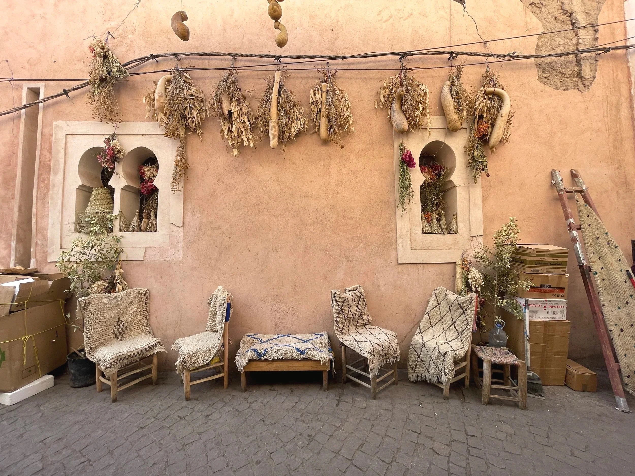 A row of chairs covered with woven fabric and blankets, placed against a peach-colored wall with arched windows and hanging dried herbs and plants, in an outdoor courtyard.