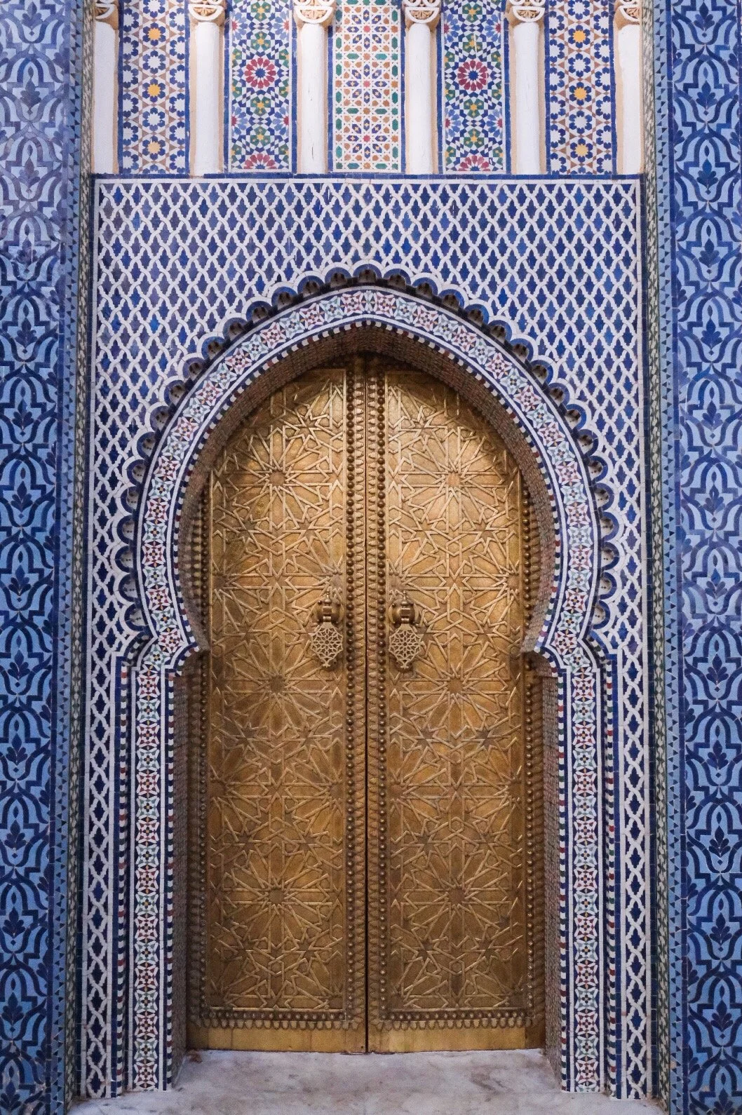 Golden metal door with intricate geometric patterns, set within an colorful mosaic arch and surrounded by blue and white tiled walls with detailed tilework.