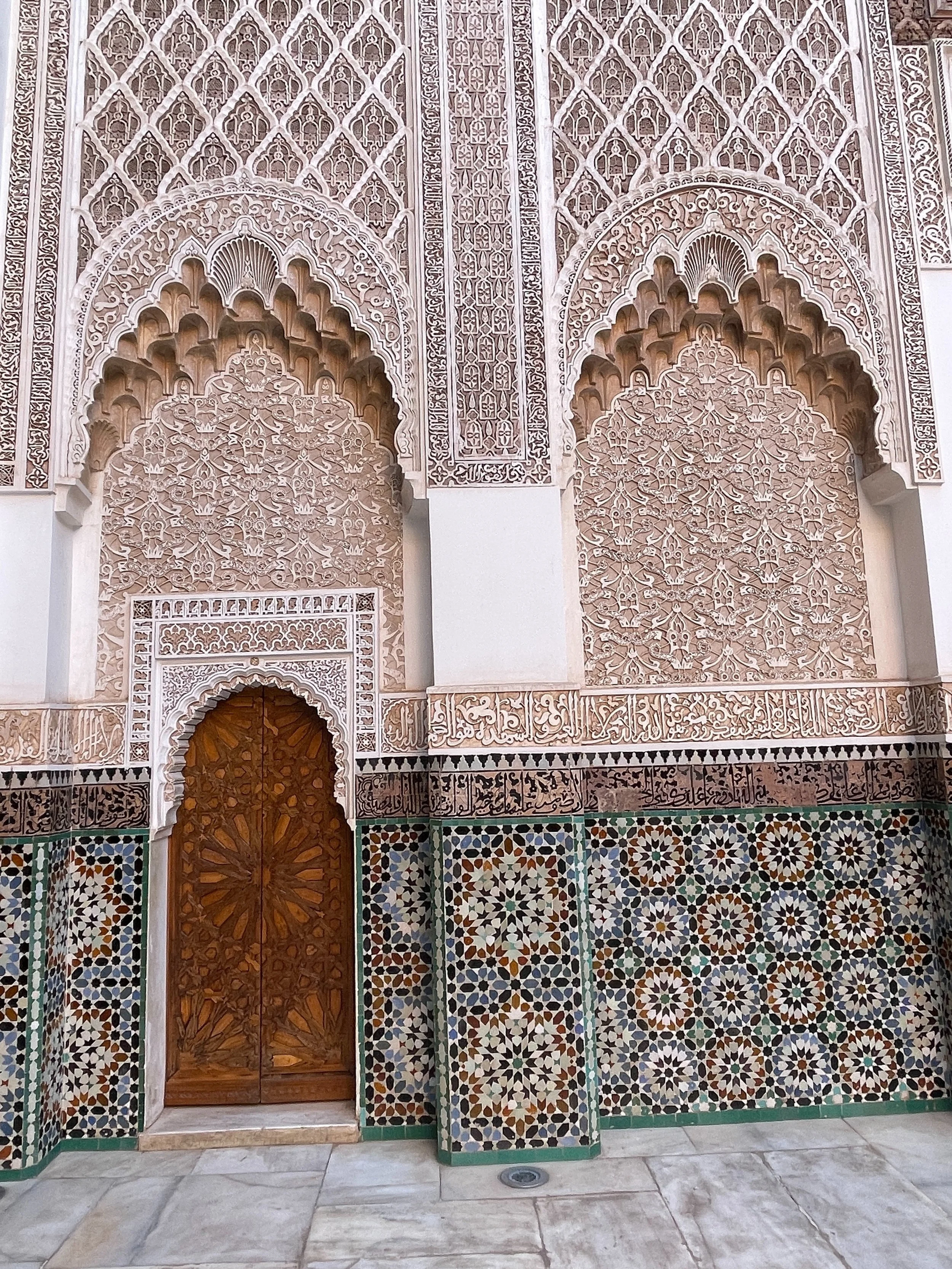 Decorative wall with intricate Islamic geometric patterns, arched door with wooden panels, and Arabic calligraphy at the bottom, possibly part of a mosque or religious building.