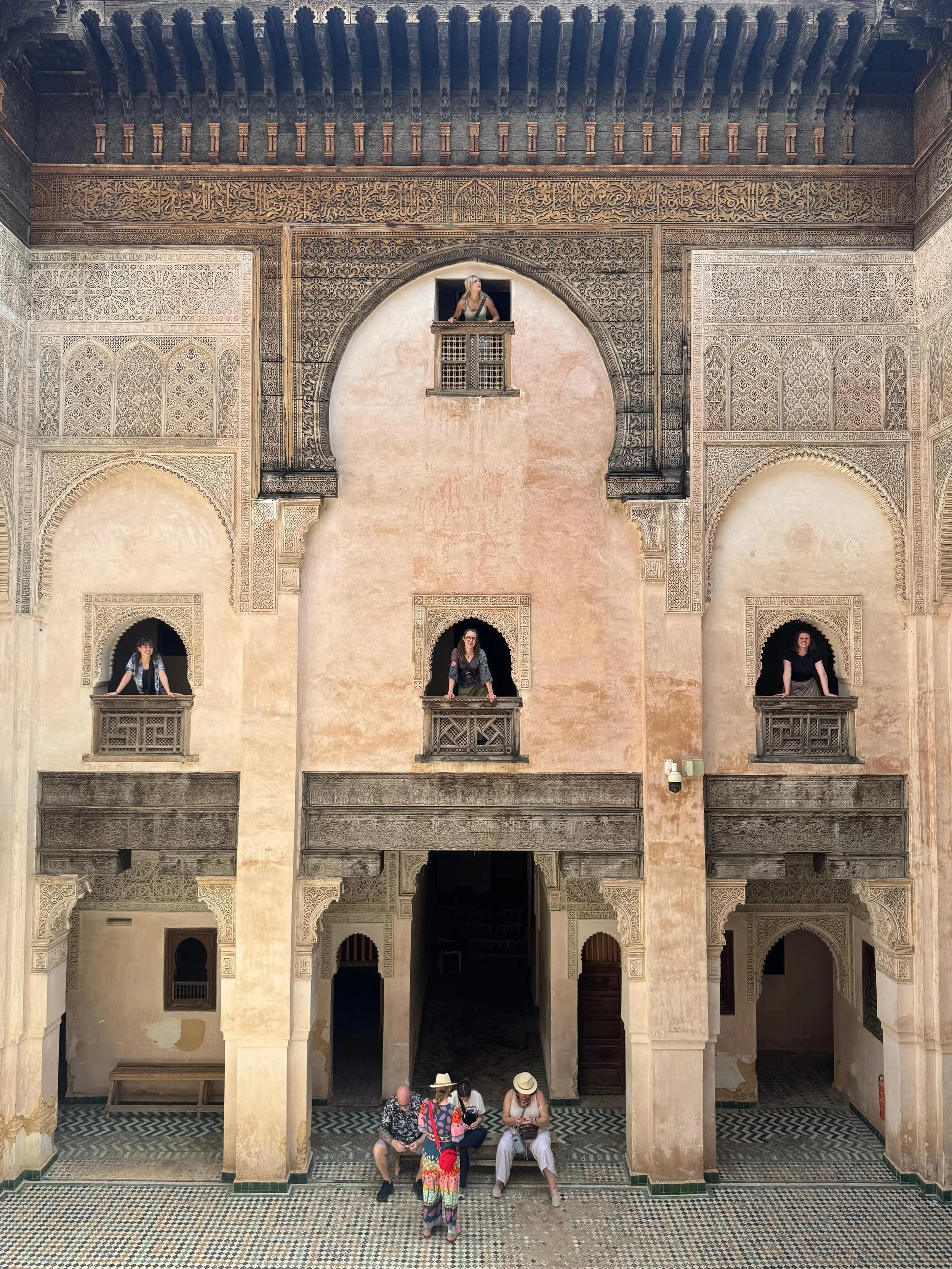 People sitting and standing in a historical building with intricate architectural details, arches, and window openings, enjoying a tour or sightseeing.