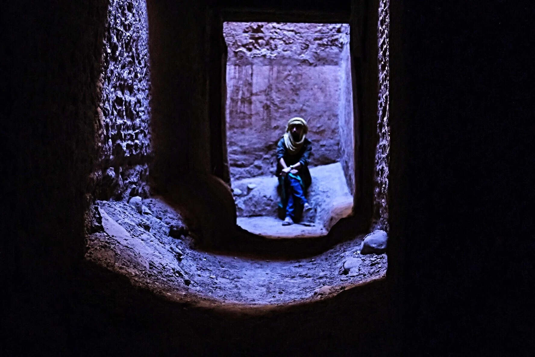 View through a small opening in a stone wall showing a person sitting against a rock wall in a cave or ancient structure, with the scene backlit by natural light.