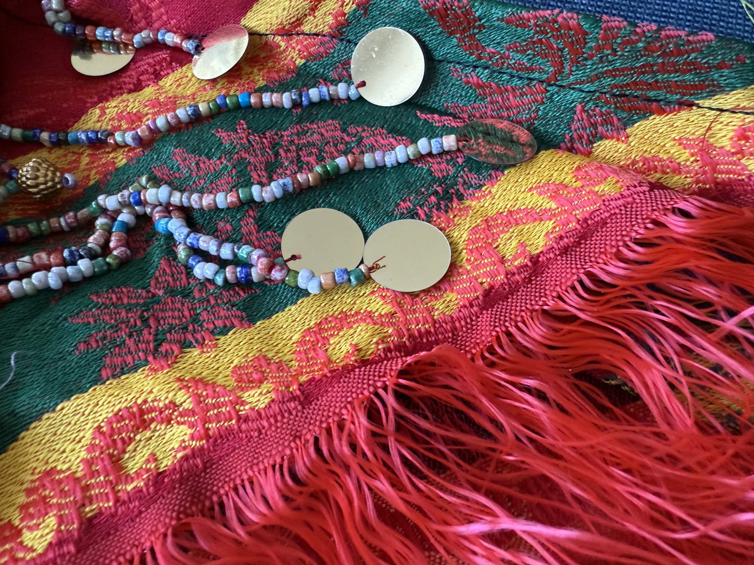 Colorful beaded necklaces with metallic round pendants laid on a vibrant woven textile with red, yellow, green, and blue patterns and fringes.