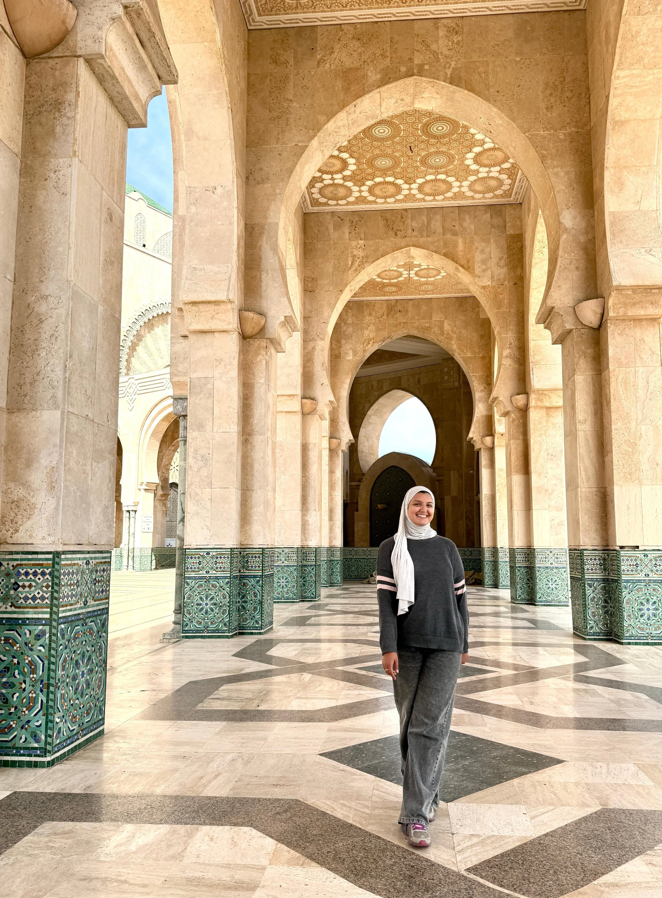 A smiling woman with a hijab standing in an ornate, arched corridor with intricate tile work, marble floors, and decorative ceiling patterns.