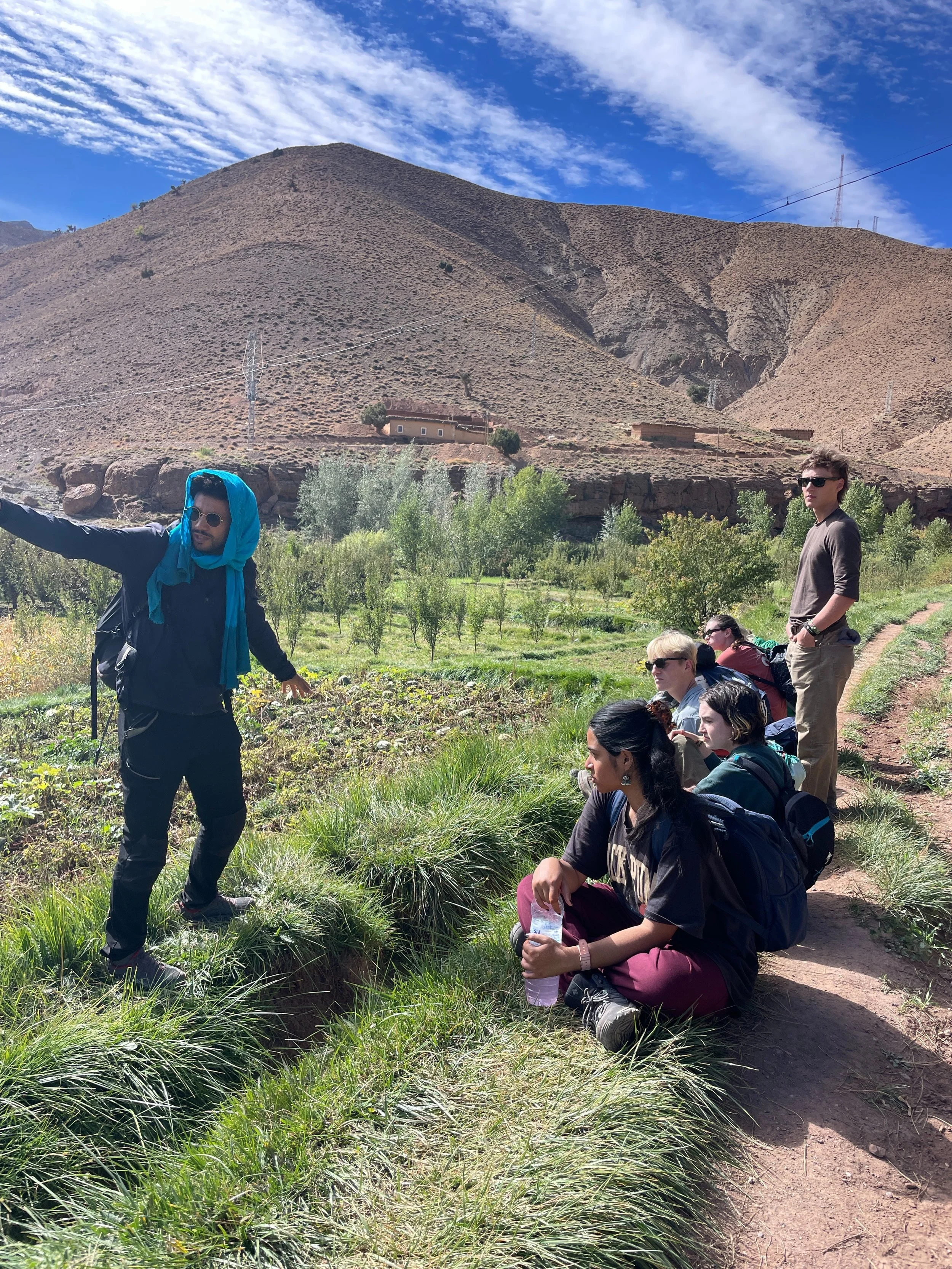 Group of hikers resting on a dirt trail in a mountainous area with sparse vegetation, with one person standing and gesturing, while others sit and stand nearby, and a dust mountain and blue sky with clouds in the background.