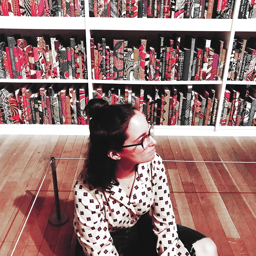 A young woman with glasses and dark hair in a bun, sitting on the floor with a patterned shirt, in front of a bookshelf filled with multi-colored fabric-patterned books.