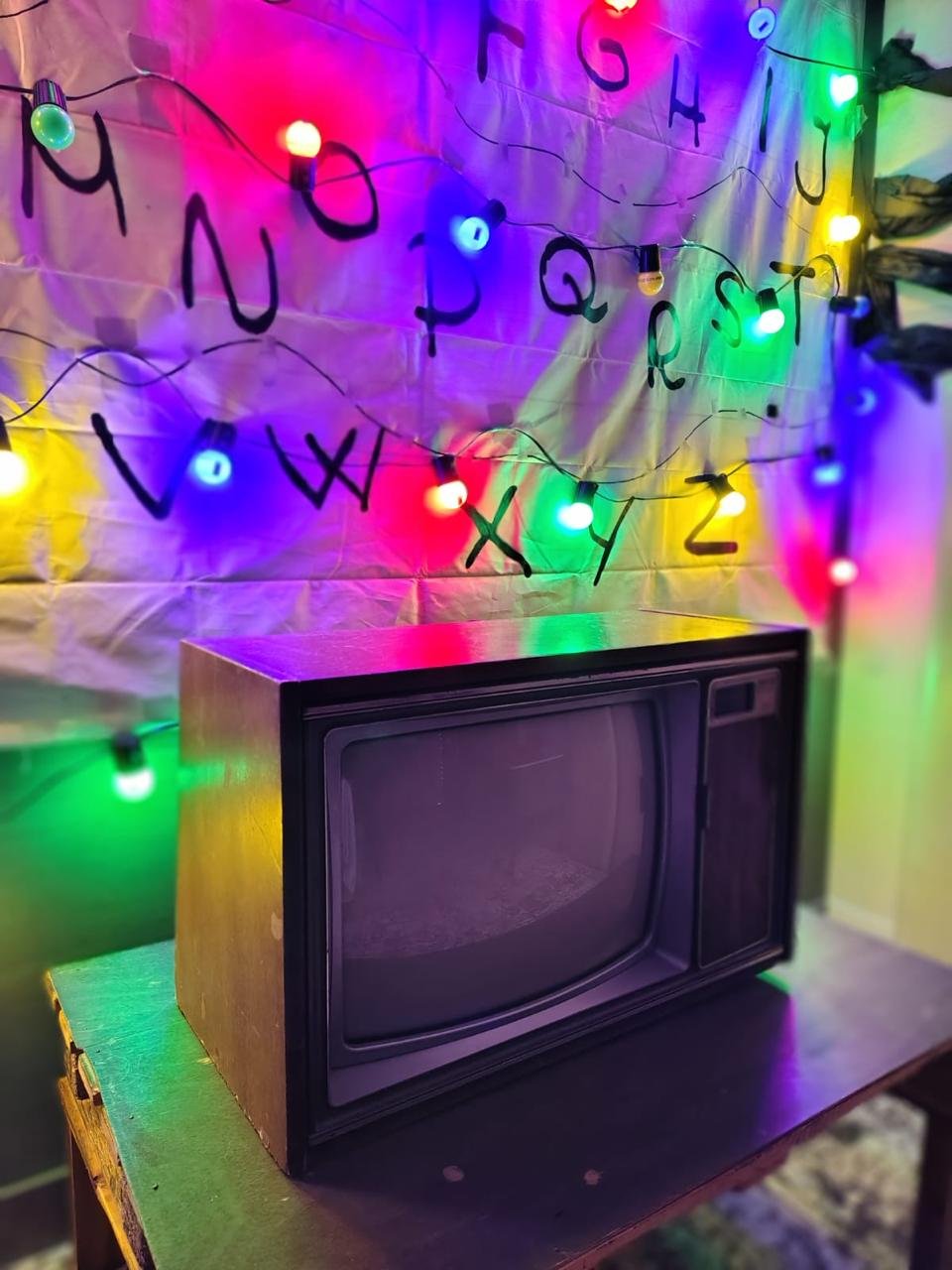Decoración con luces de colores y letras del abecedario colgando en una pared con telarañas artificiales y un televisor antiguo en una mesa.