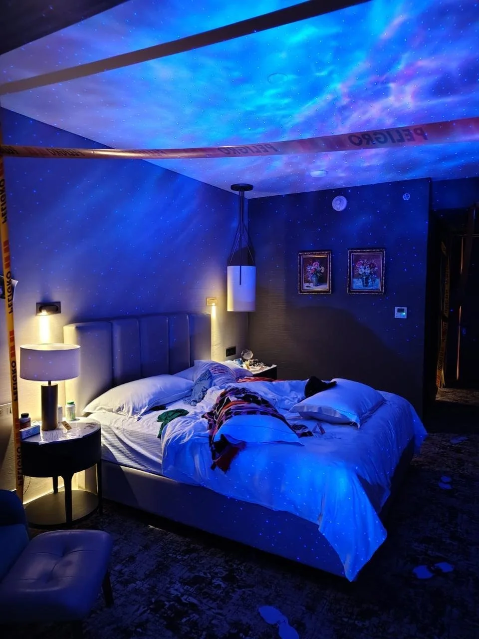 Habitación con proyección de estrellas y galaxias en el techo, creando un efecto de cielo estrellado, con cama desordenada, lámparas encendidas y cuadros en la pared.