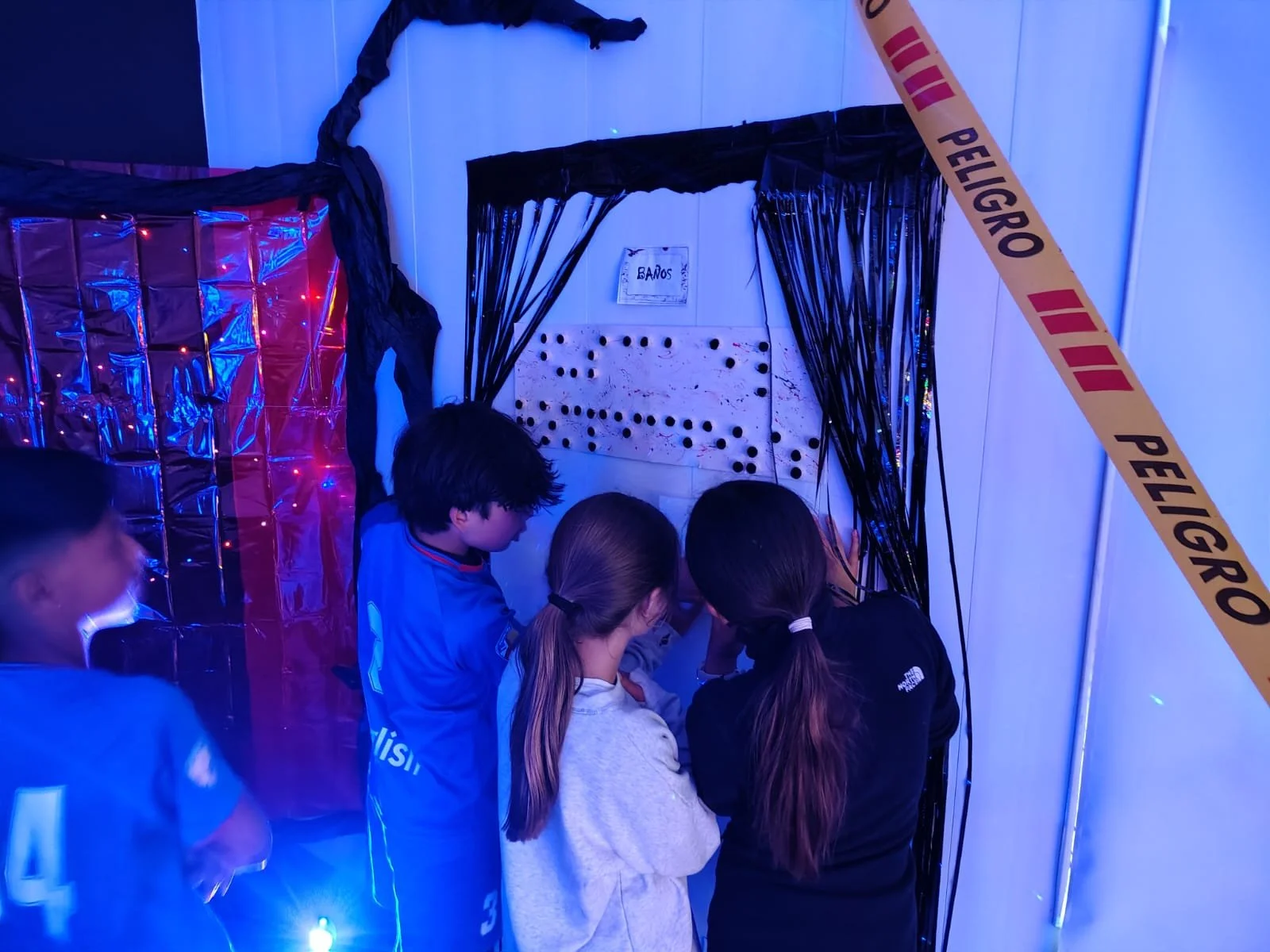 Niños observando una pared con agujeros en un espacio decorado con luces de colores y cintas negras, en una zona marcada como 'Baños' con una cinta de advertencia naranja.