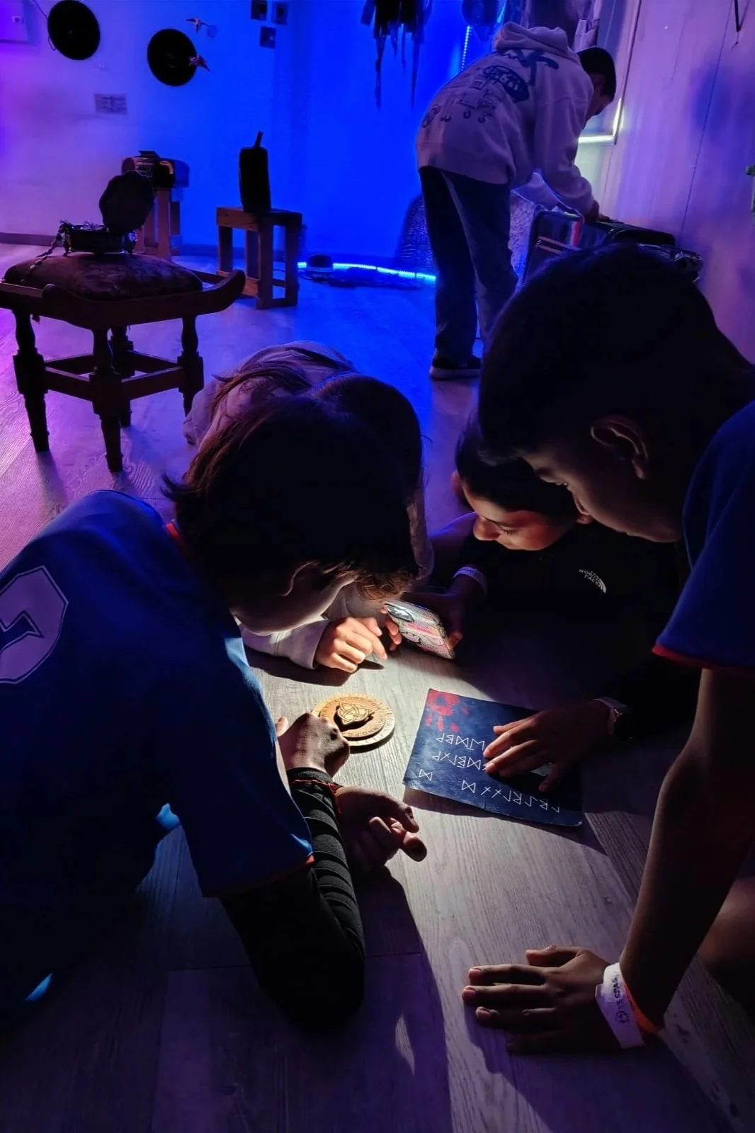 Niños en un grupo mirando un teléfono móvil en un ambiente oscuro con iluminación azul, en el suelo y con objetos de estilo futurista.