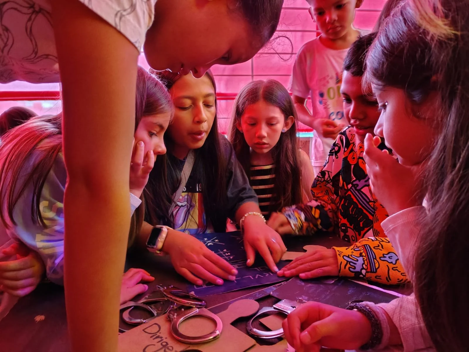 Grupo de niños y una mujer observando y aprendiendo cómo usar tijeras y otros instrumentos en una mesa, en un entorno con iluminación de color rosa.